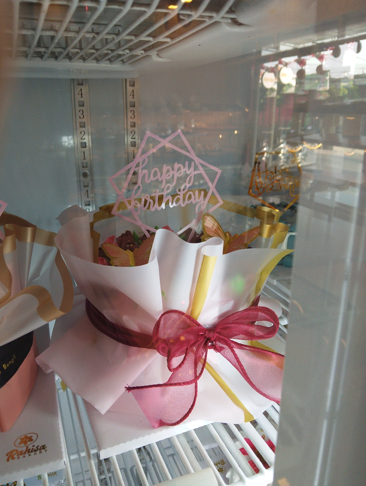 In Manaruwi, Indonesia, a meticulously arranged bouquet of pastries rests within a refrigerated display case. The bouquet, wrapped in delicate white and gold paper, is adorned with a vibrant pink "Happy Birthday" topper and a crimson ribbon tied into a bow. The pastries themselves are artfully decorated with floral accents and a charming butterfly. The background reveals the interior of the display case with numbered shelf guides and the soft glow of ambient lighting. The image captures a festive and delicious offering, ready to be enjoyed.