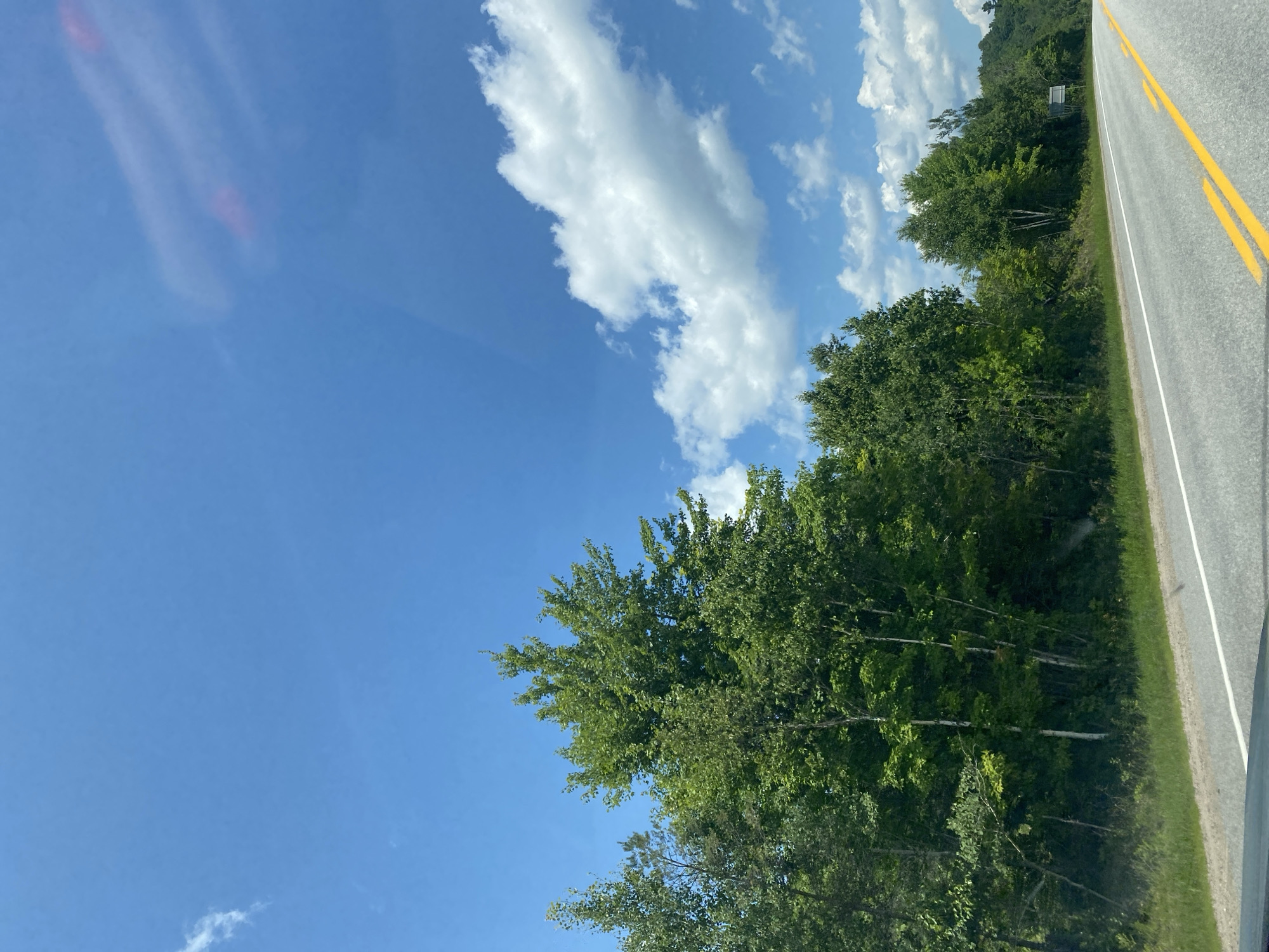 The image depicts a roadside view of a highway in Chénéville, Canada, under a bright blue sky with scattered white clouds. The highway, paved with asphalt, features a solid white line and double yellow lines marking the lanes. To the left of the road, a dense line of green trees and foliage forms a natural barrier. The trees are lush and varied in height, with some taller specimens reaching towards the sky. The ground alongside the road is covered with green grass. In the distance, the terrain appears to be gently rolling hills also covered in trees. A road sign is visible to the far right of the image, partially obscured by trees. The overall impression is a serene, natural landscape on a clear day.