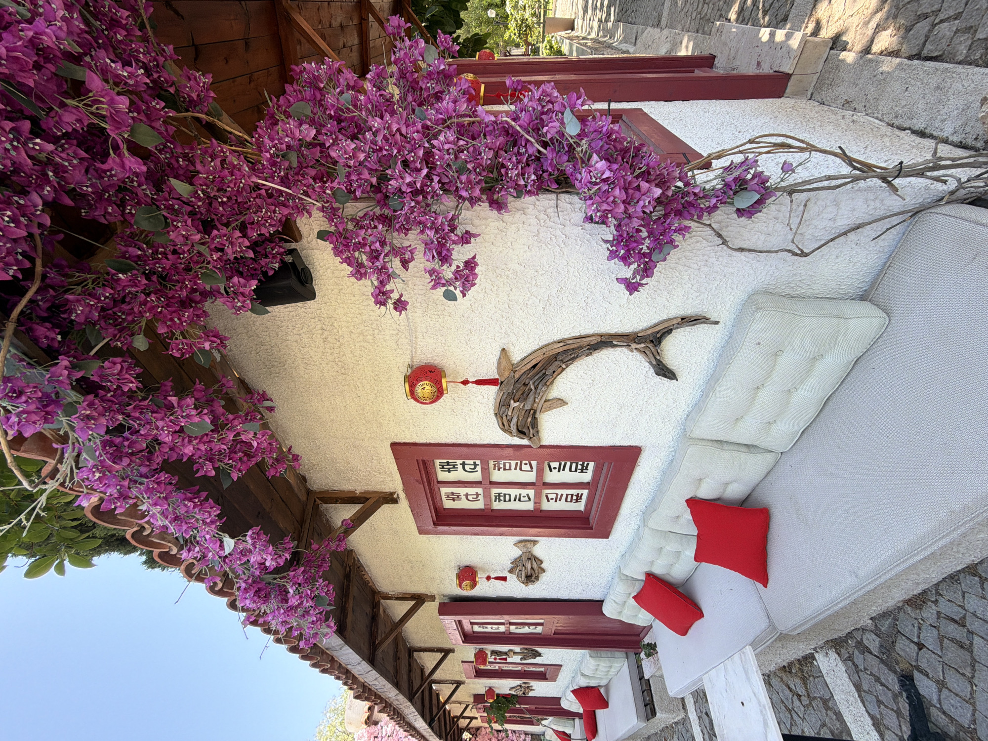 The image depicts a charming exterior scene, likely a cafe or restaurant, in Kemer, Turkey. The building features a textured white stucco facade with red-trimmed windows and doors, some of which are open. Bougainvillea flowers in a vibrant magenta hue cascade over the roofline and down the side of the building, adding a burst of color.
Along the exterior wall, there are two windows and two doors visible. The windows have red frames and feature a decorative panel with what appear to be Asian characters. Red Chinese lanterns are hung above the windows and doors, complementing the red accents of the building. A wooden sculpture of a dolphin is mounted on the wall between two of the windows.
In front of the building, a long, low-profile sofa with light-colored cushions and bright red accent pillows is positioned along the wall. The sofa sits on a stone base, and a small white marble-topped table is placed to the left of the leftmost seating area. The ground is paved with irregular cobblestones.
The scene is bathed in bright sunlight, suggesting a clear day. The overall impression is one of a welcoming and aesthetically pleasing outdoor space, typical of a resort town like Kemer.