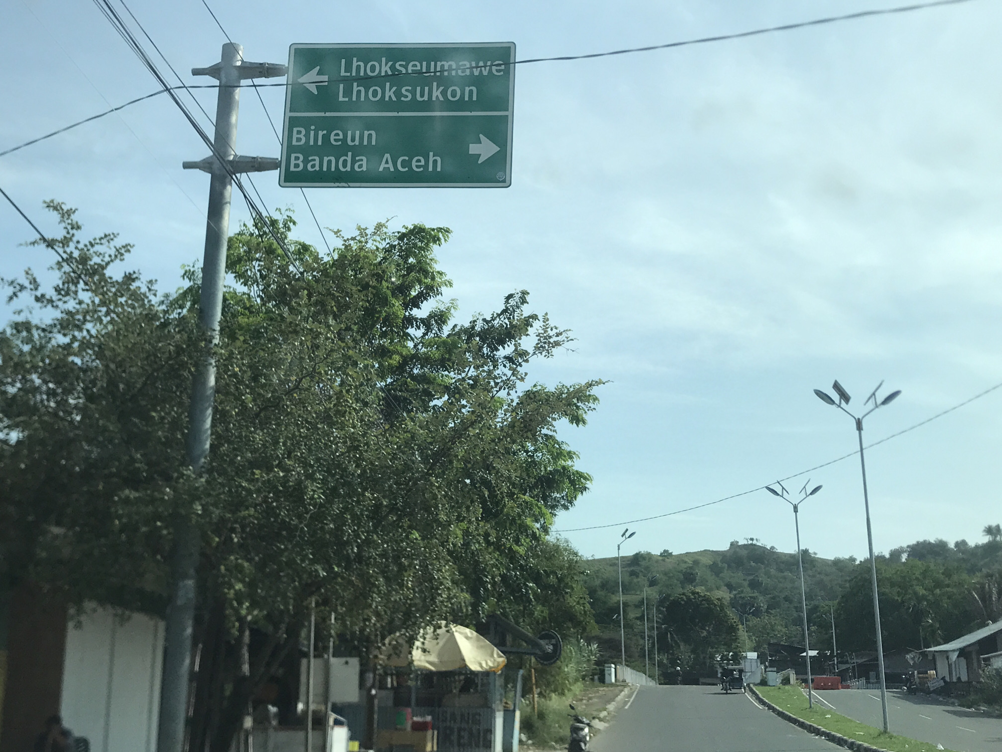 The image captures a bright, daytime roadside scene in Lhokseumawe, Indonesia, likely from the perspective of a vehicle. A prominent green directional road sign, mounted on a grey utility pole, dominates the upper left side.
The sign indicates directions to multiple locations: "Lhokseumawe" and "Lhoksukon" are pointed to the left by an arrow. Below these, "Bireun" and "Banda Aceh" are indicated with a right-pointing arrow. The utility pole supports several overhead power lines against a clear, partly cloudy sky.
Lush green trees line the left side of the road, partially obscuring a building. On the right, a small street vendor stall with a light-colored, ruffled umbrella is visible, next to which a black scooter is parked. Partial text on the stall reads "ORGANIC DURENG". The asphalt road features a low concrete median strip and extends into the distance, bordered by modern streetlights. A verdant, undulating hill with scattered structures forms the background. The scene suggests good weather and a typical bustling roadside environment in a tropical region.