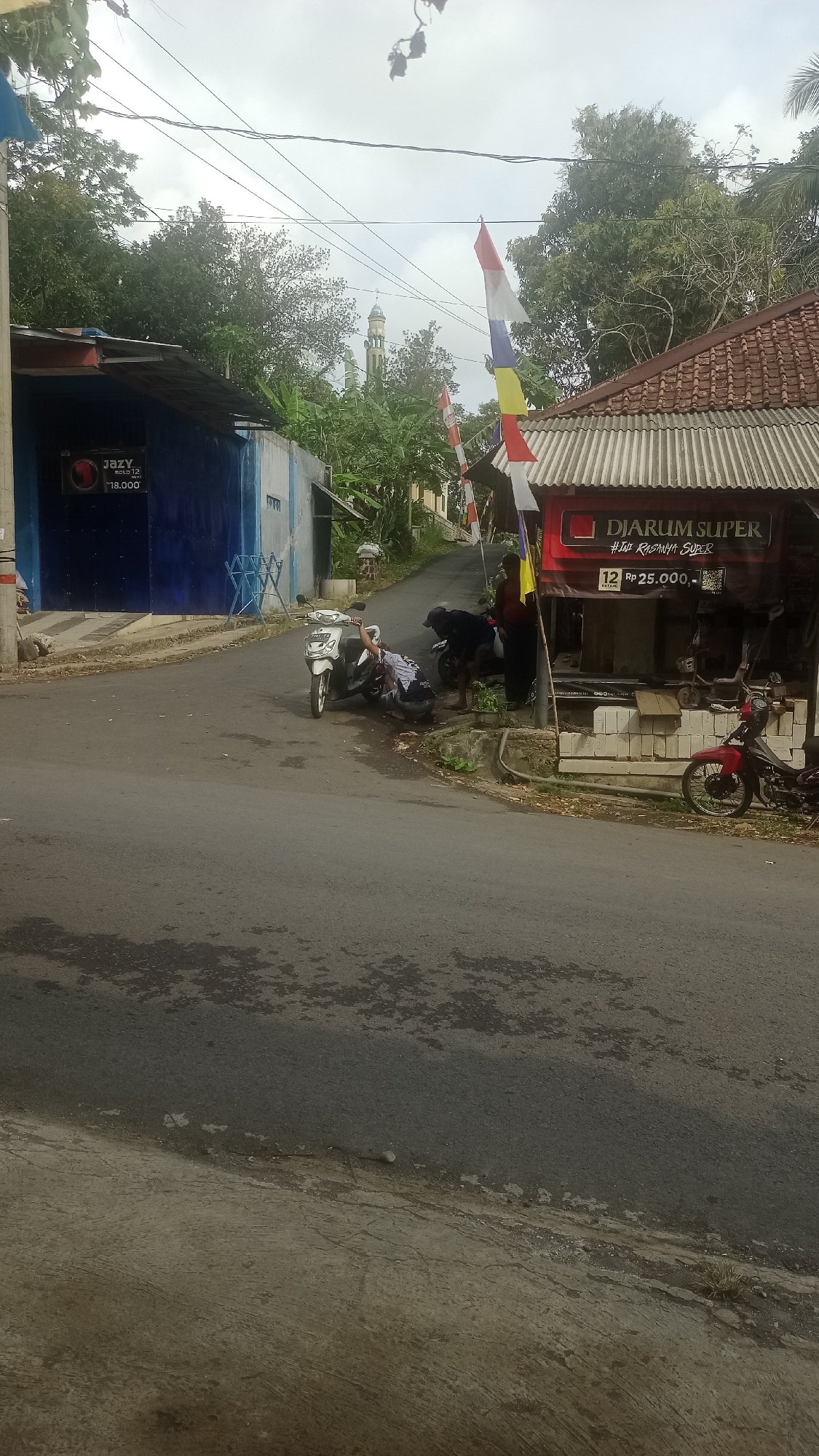 The image captures a street scene in Karyamandala, Indonesia. On the left, a blue building with a sign that reads "JAZY moto 12.000" is visible. To the right, a building with a red sign stating "DIARUM SUPER #JADI RASANYA SUPER 12 RP 25.000" is present. A white scooter is parked on the street, with one person leaning over it. Another scooter, red in color, is parked further down the street. A person in dark clothing is standing near the red building. The street is paved, with some wet patches. Trees and a mosque minaret are visible in the background. Several Indonesian flags are displayed, fluttering in the wind.