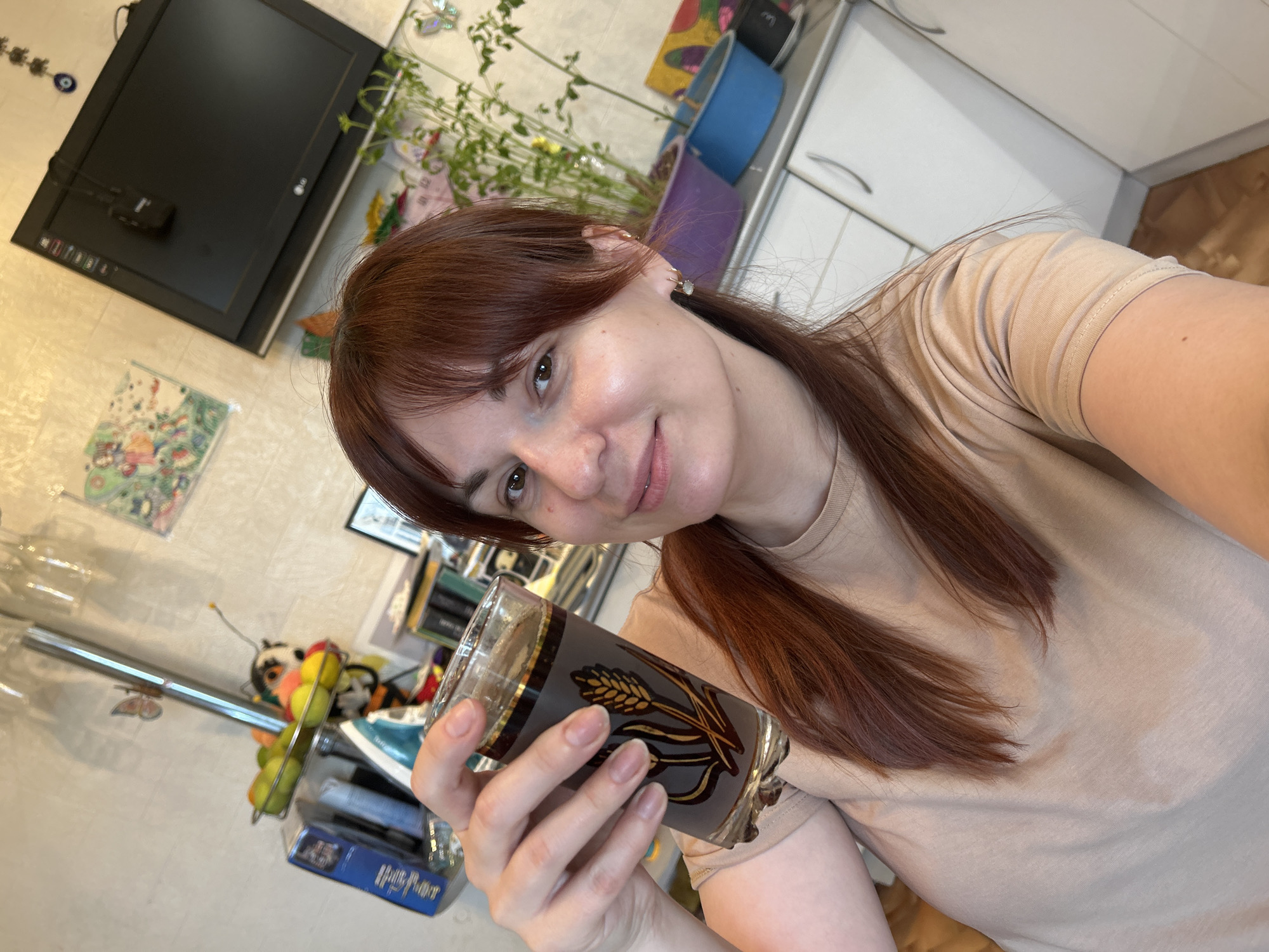 A woman is holding a glass filled with a dark liquid. The glass has a wheat stalk design. She is wearing a beige t-shirt and gold earrings. The background includes a kitchen with white cabinets, a black television, a fruit basket, and a plant. The location appears to be an indoor setting in Фоки, Russia.