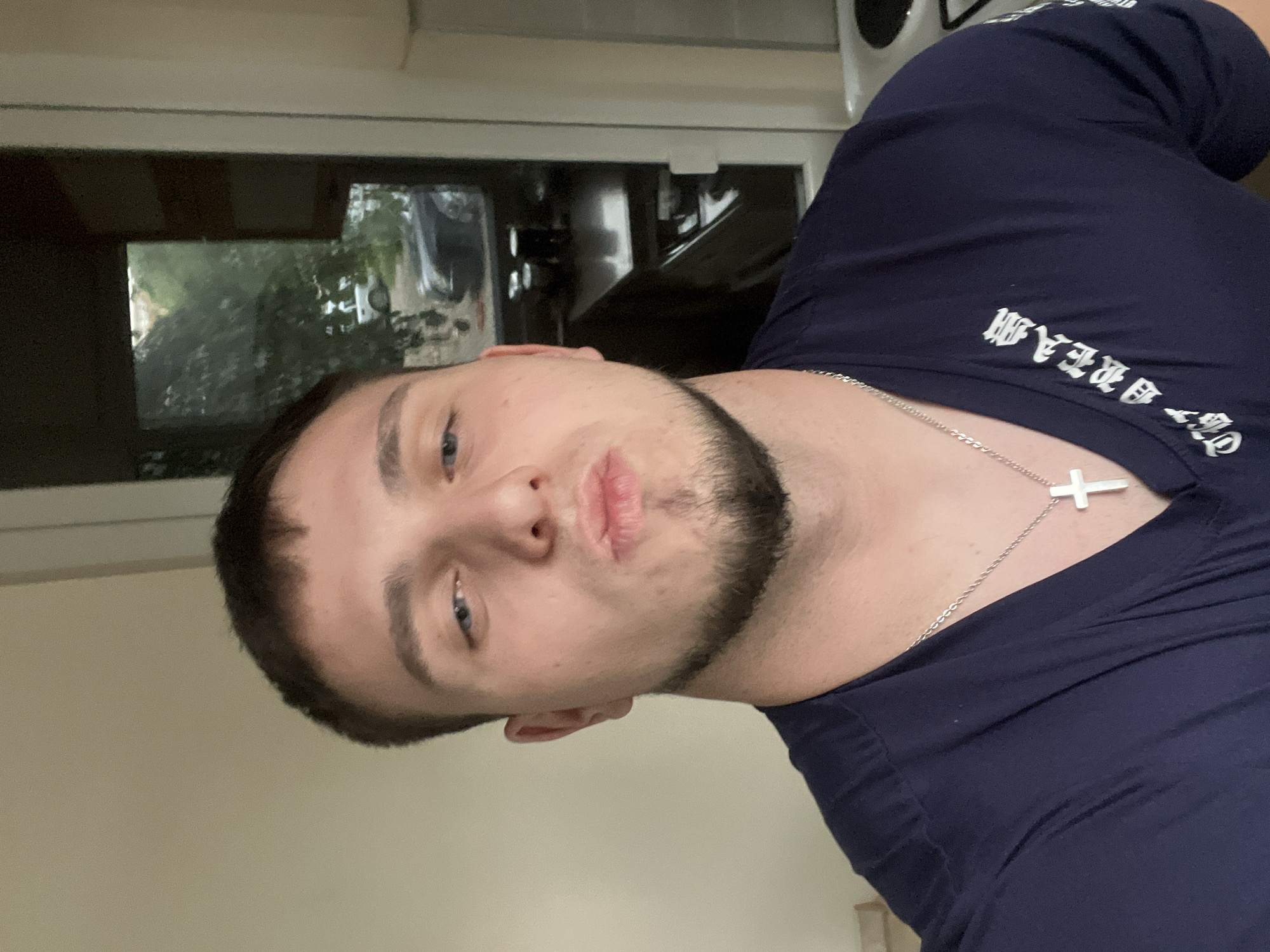 A close-up, tilted, selfie-style photograph shows a young, fair-skinned male with short dark hair and a beard. He is looking directly at the camera with a slightly pursed-lip expression. He is wearing a dark blue t-shirt with white gothic-style text on the upper chest, and a silver chain necklace with a cross pendant. The background shows an interior space with a window. Reflections in the window suggest an outdoor environment. Specific location information in Plovdiv, Bulgaria is unavailable.