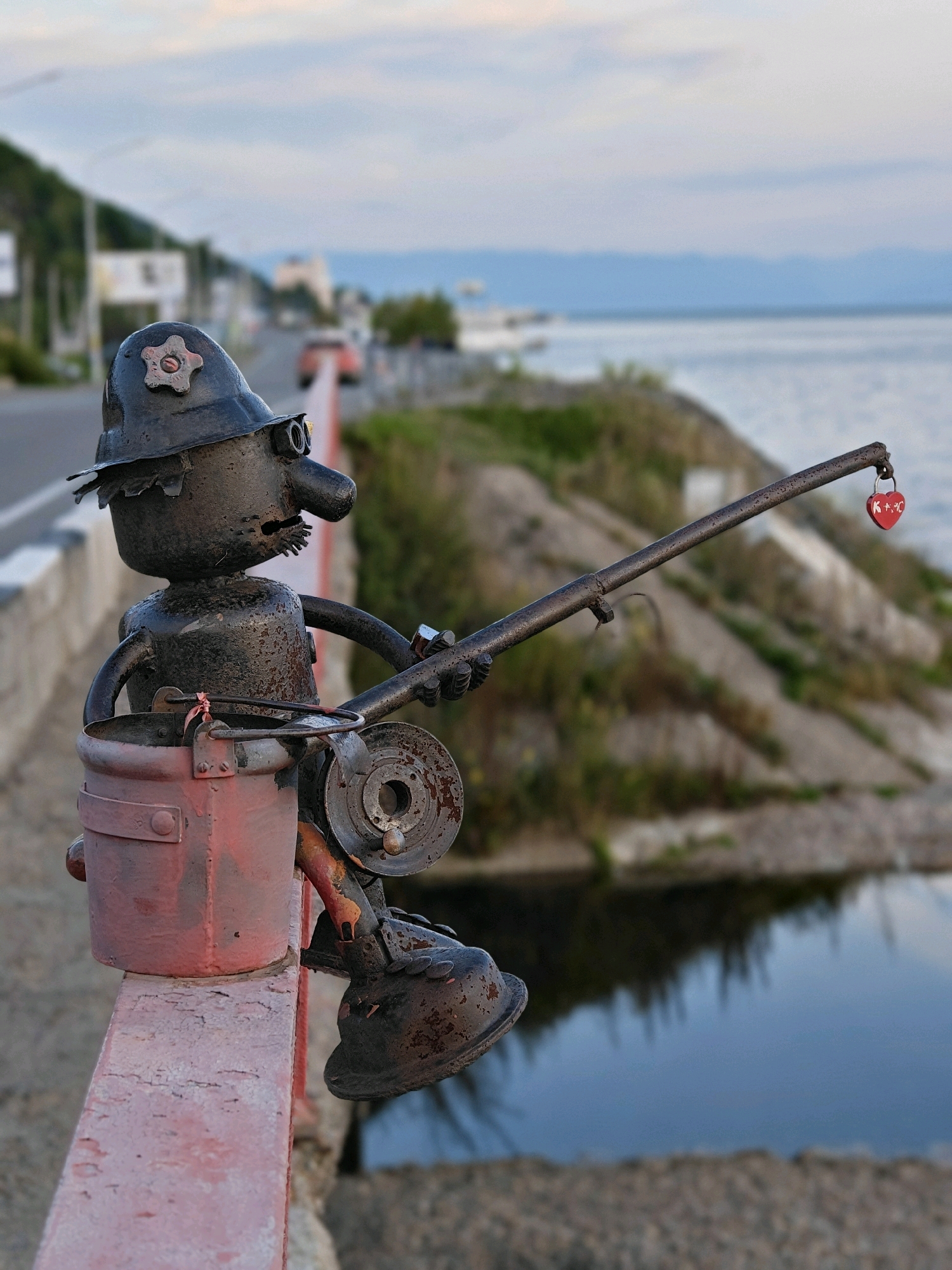 An iron sculpture of a fisherman, made from scrap metal, sits on a red railing overlooking a body of water. The fisherman is depicted wearing a hat and goggles, holding a fishing rod with a heart-shaped padlock attached to the end. A bucket hangs from the fisherman's arm. In the background, a road runs alongside the water, with trees and buildings visible further in the distance. The sky is overcast, with a soft light suggesting it is either early morning or late evening. This sculpture is located in the city of Listvyanka, Russia, a popular tourist destination on the shores of Lake Baikal.