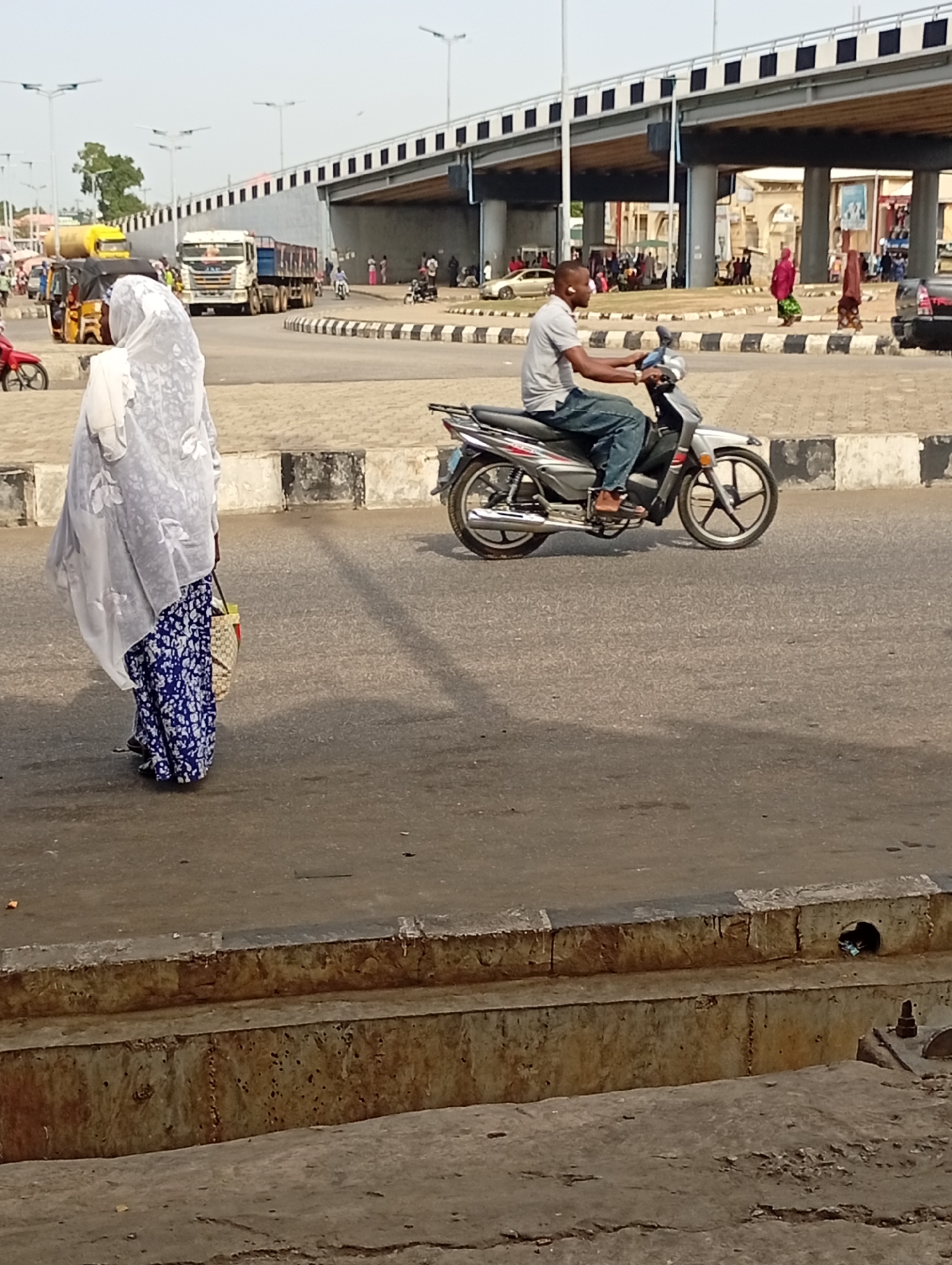 The photo was taken in Lokoja, Nigeria. A woman wearing a white head covering and a long blue floral skirt stands on the side of a road. Next to her is a concrete curb. A man on a silver motorcycle is riding on the road, facing right. Behind him, there is a bridge. Many people are standing and walking around under the bridge. There are cars and trucks on the road behind the bridge.