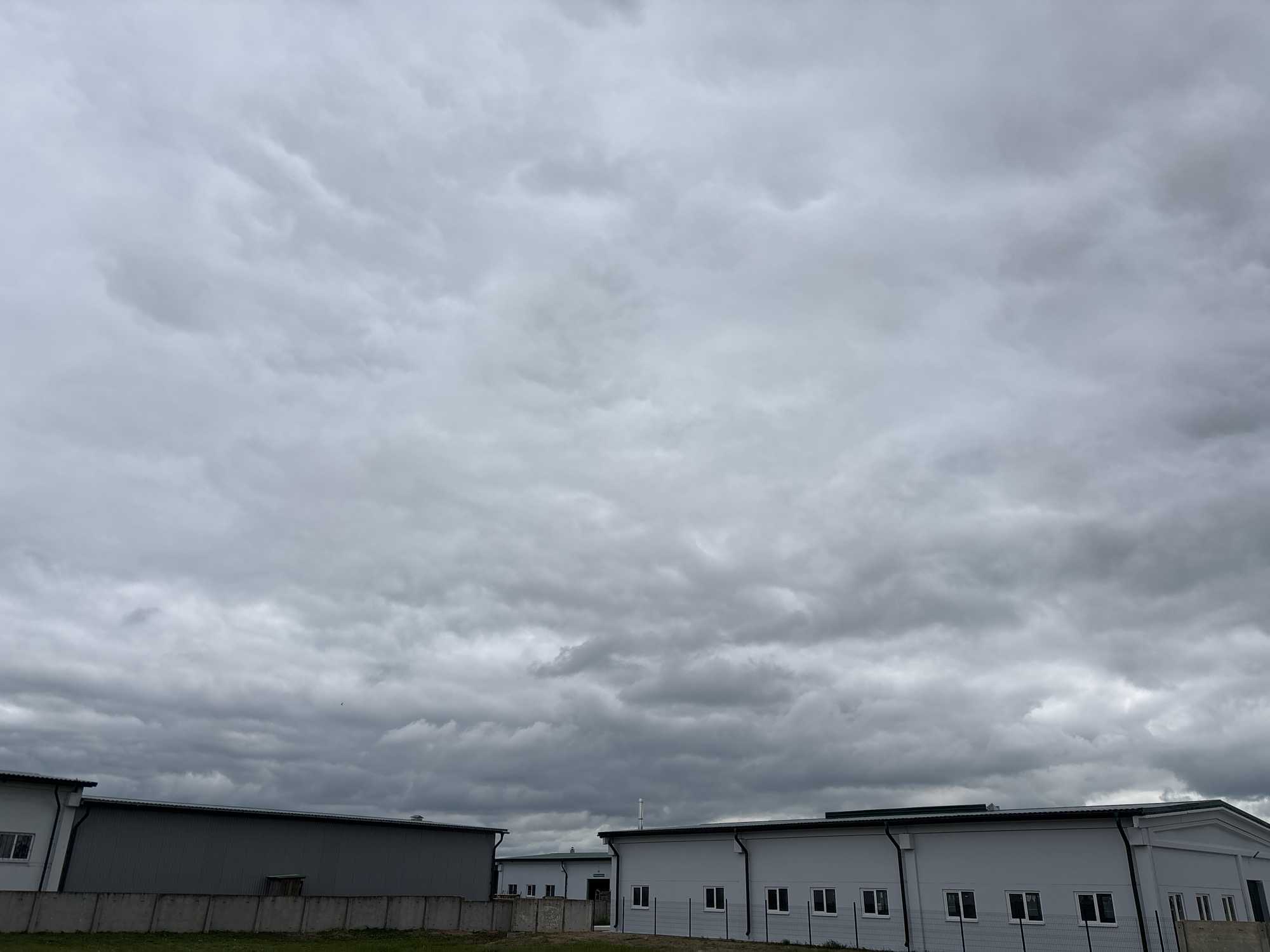 A wide-angle, eye-level shot captures a cluster of modern industrial buildings under a heavily overcast, grey sky. The buildings, primarily white with dark grey roofs, feature numerous rectangular windows and clean lines. In the foreground, a low concrete fence with a wire mesh top separates the buildings from a strip of green grass. To the left, a larger grey building with a flat roof is partially visible, with a section of darker grey cladding. The sky dominates the upper two-thirds of the image, filled with dense, textured grey clouds that suggest an impending or recent rainfall. A single small dark object, possibly a bird, is visible in the sky above the buildings. The scene appears to be located in an industrial area of Капыльскі сельскі Савет, Belarus.