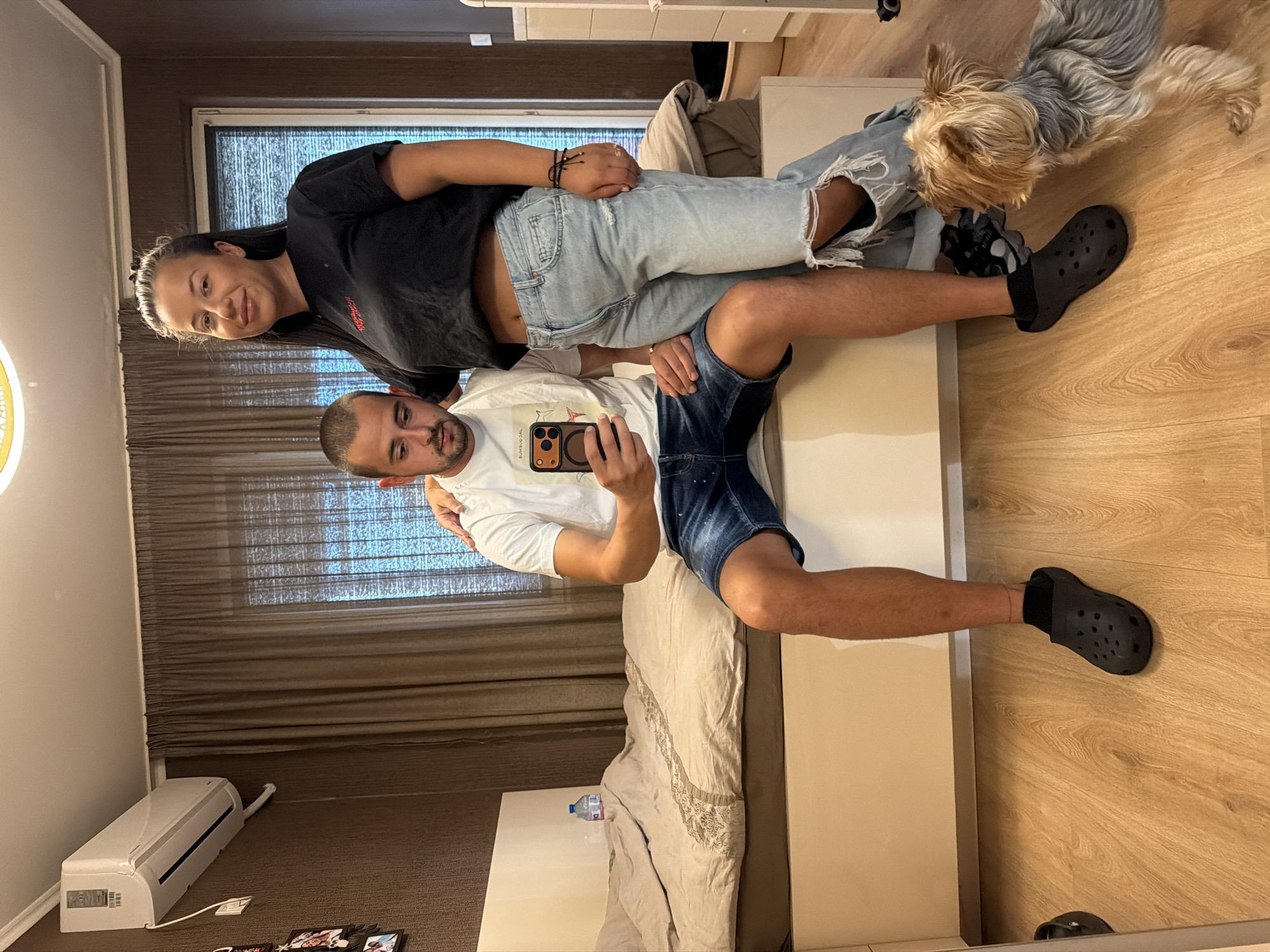 An adult male and female are posing for a selfie in a bedroom. The male is sitting on the edge of a bed, holding a smartphone, and wearing a white t-shirt and denim shorts. The female is standing behind him, with her hand on his shoulder, wearing a black t-shirt and ripped jeans. A small dog is looking at the couple from the floor. The room has a bed with beige bedding, a dark brown curtain, and an air conditioner mounted on the wall. The floor is made of light-colored wood. The overall scene appears to be a casual, indoor setting. The location of Burgas, Bulgaria, cannot be determined from the visual information provided in the image.