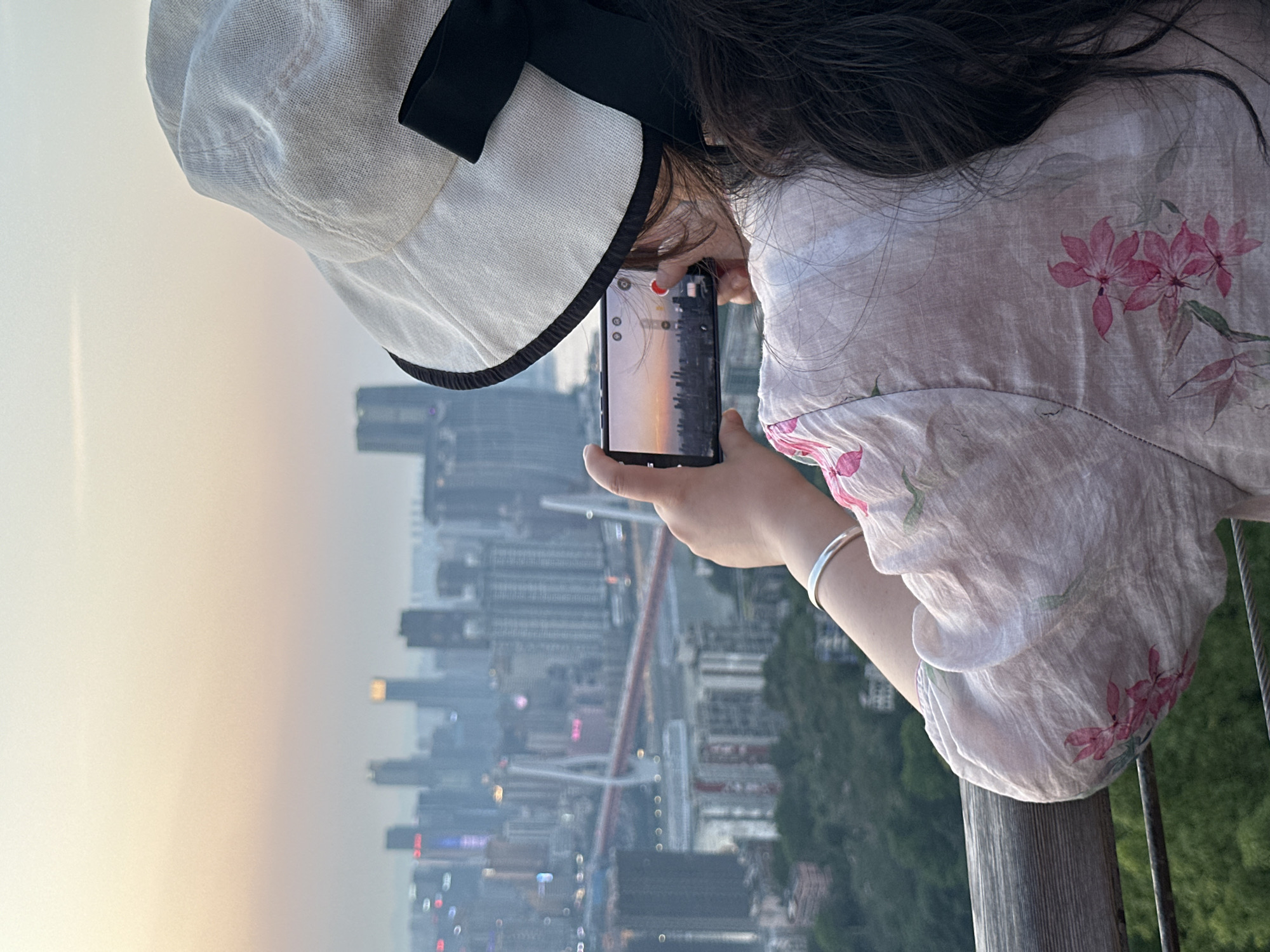 A person with dark hair and a white hat with a black ribbon is seen from behind, holding a smartphone to take a video or photo of a cityscape. The cityscape includes tall buildings and bridges, likely taken from a viewpoint in Nan'an District, China. The person is wearing a floral-patterned top and a silver bracelet. They are leaning against a wooden railing.