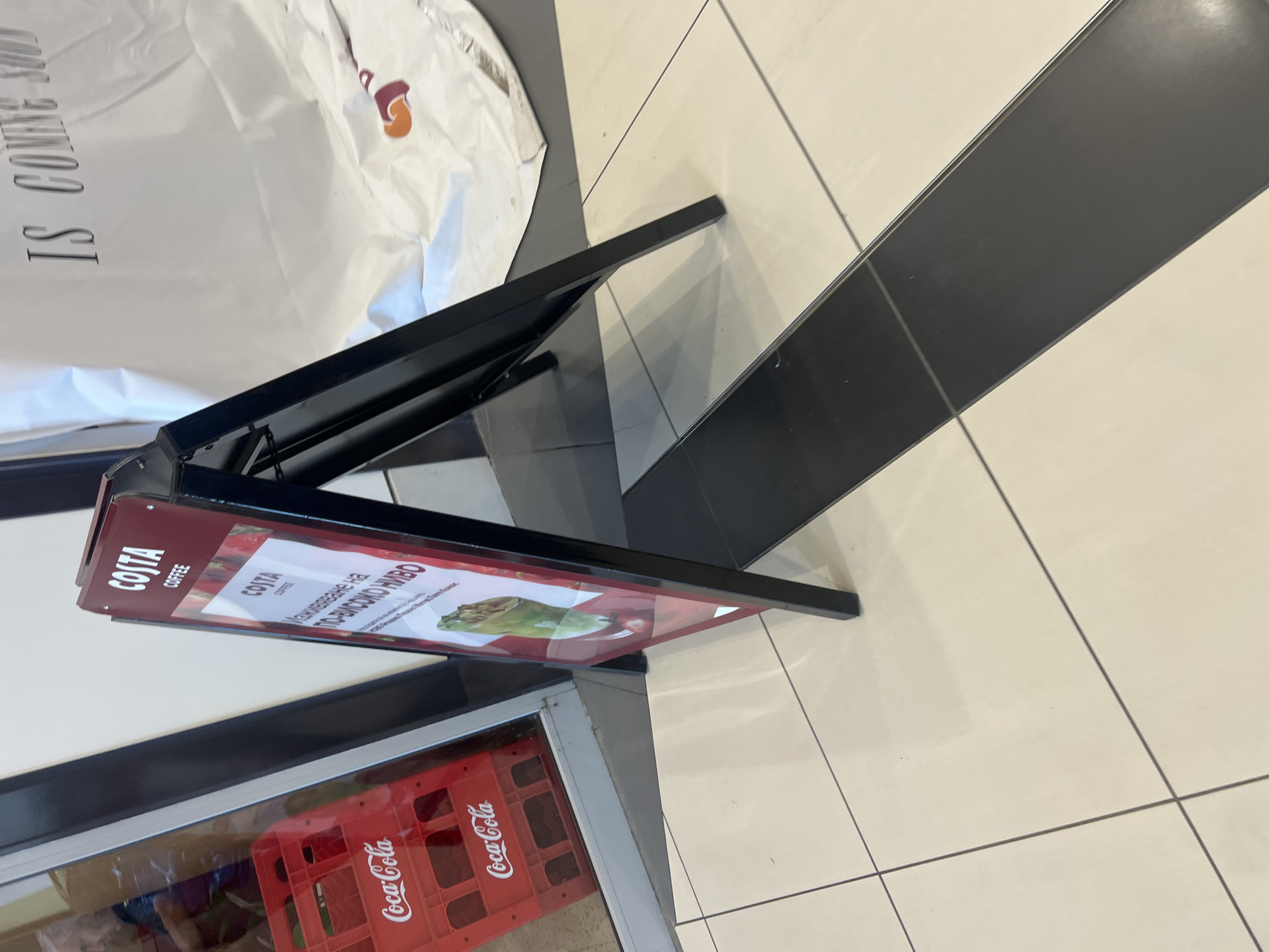 The image depicts a Costa Coffee advertising sign standing near a shop window. The sign is an A-frame structure with the Costa Coffee logo and promotional text in Bulgarian, reading "Изживяване на по-високо ниво," which translates to "Experience at a higher level." The sign also features an image of a Costa Coffee product. Through the shop window, a red Coca-Cola crate is visible. The location appears to be indoors on a tiled floor. Further details about people, subjects, objects, actions, interactions, and the environment:
- No humans are visible in the image.
- The sign suggests the presence of a Costa Coffee establishment nearby.
- The environment is a commercial space with a tiled floor.
- The lighting is bright and uniform.
- The angle of the photograph suggests it was taken from a standing position.
- The overall impression is of a commercial environment.
Burgas, Bulgaria, is not mentioned.