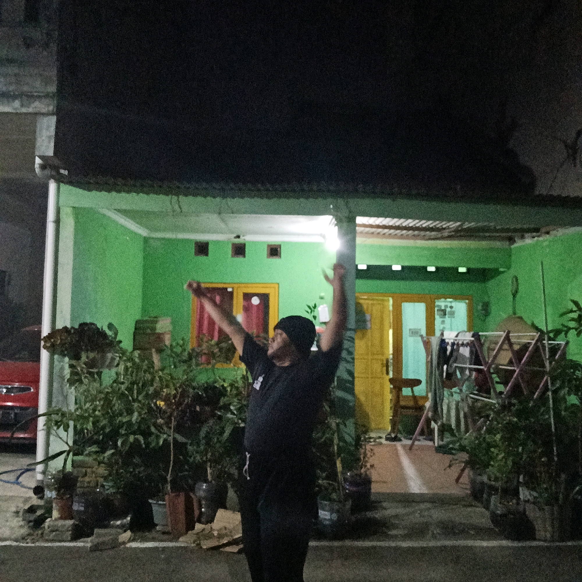 A person wearing a black t-shirt and black beanie stands in front of a bright green house at night, with their arms raised and a slight smile on their face. The house has yellow doors and windows with red curtains. Potted plants are arranged in front of the house, and a red car is partially visible on the left. A clothesline with laundry is set up on the right side of the house. The scene appears to be in a residential area in Salatiga, Indonesia, given the architectural style and the presence of potted plants common in Indonesian homes.