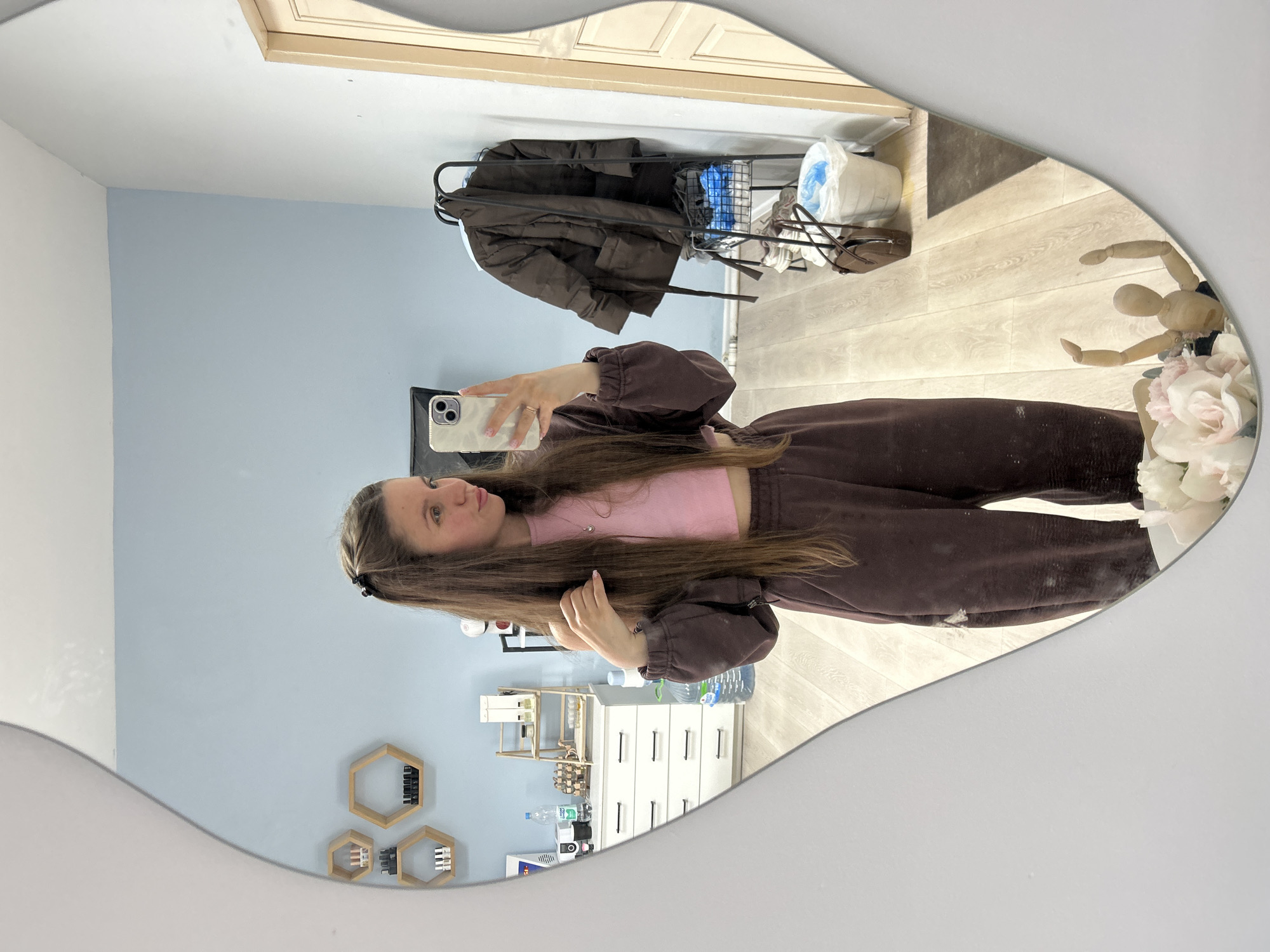 A young woman with long brown hair is taking a selfie in a mirror. She is wearing a pink top and brown sweat suit. The background includes a light blue wall with hexagonal shelves, a white dresser with shelves, a coat rack with a brown jacket, and wooden floors. The location is likely an apartment in Klin, Russia, based on the user query. There's also a decorative wooden figure and flowers visible in the mirror.