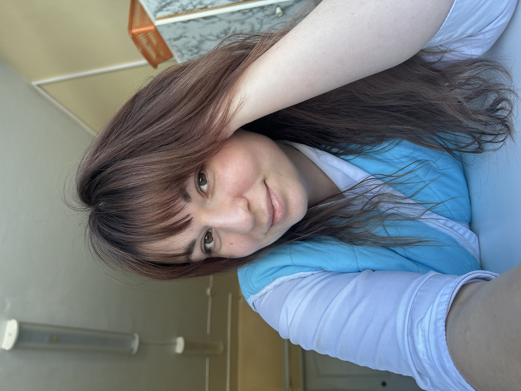 A female subject, approximately 20-35 years of age, with medium-brown hair featuring reddish-brown highlights and bangs, looks directly at the camera with a slight smile. She has brown eyes and a light skin tone. Her left arm rests on a white horizontal surface in the foreground, while her right arm is positioned behind her head, with the elbow visible against her long hair. She wears a light blue quilted vest over a white collared shirt. The background reveals an indoor setting: a light-colored ceiling with a long, white cylindrical fluorescent light fixture above the subject's left side. To the right, a light yellow wall with white trim molding is visible, along with an orange perforated plastic basket resting on a grey and white marbled surface, possibly a cabinet or countertop. A light-colored door or cabinet is partially visible in the left background. The scene is located in the city of Фоки, Russia.