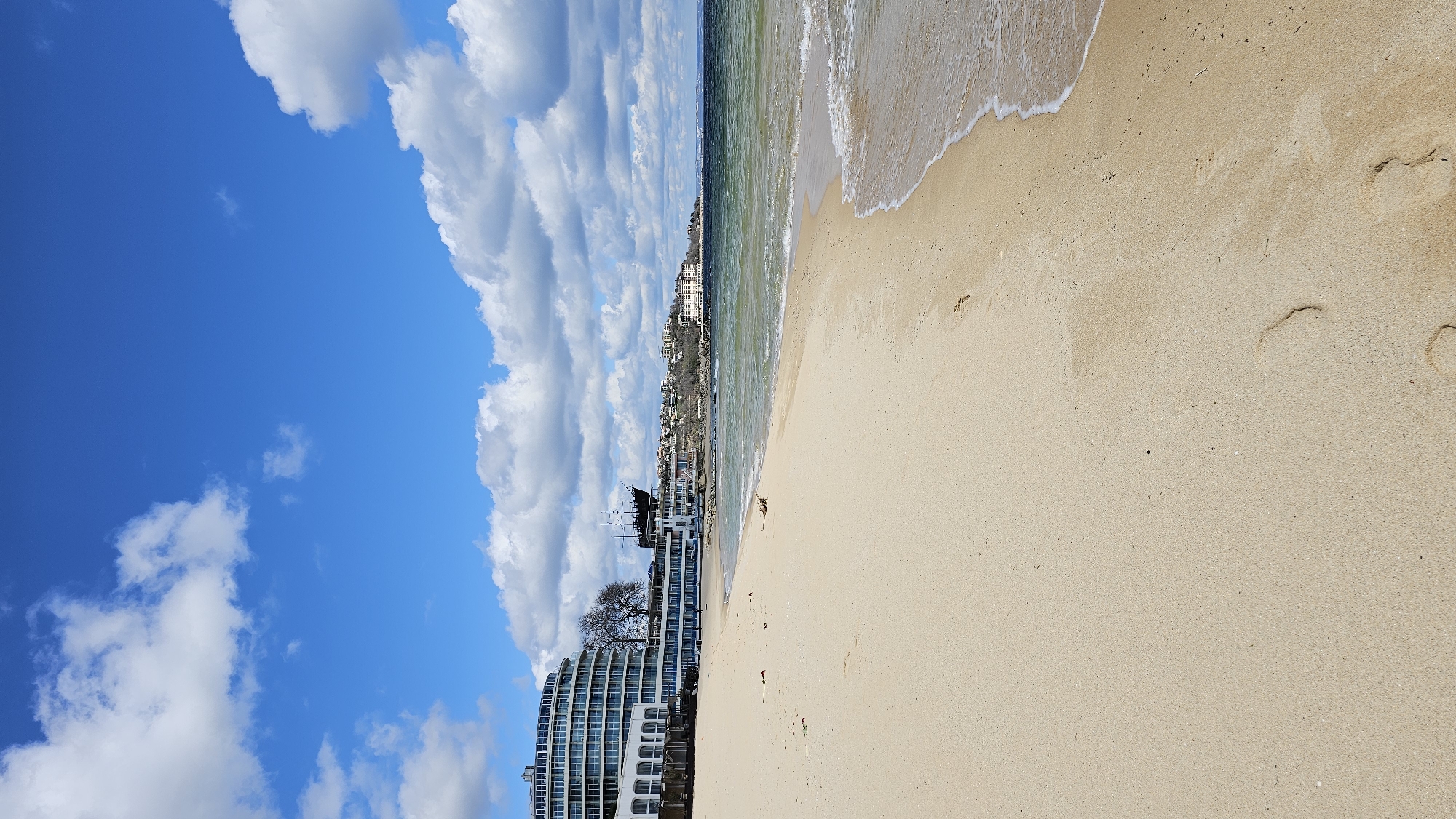 The image shows a beach in Varna, Bulgaria. The view includes a sandy beach, the sea, and a blue sky with cumulus clouds. The shoreline has gentle waves. Buildings, including a tall modern hotel with a curved facade, are visible in the background. A pirate ship replica is located near the water and hotel. Footprints are visible in the sand.