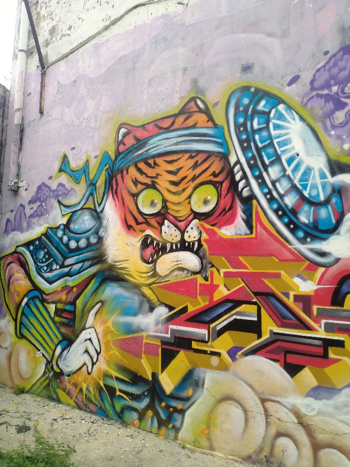 This full-shot image captures a vibrant graffiti mural on a weathered light purple wall, likely in an urban outdoor setting in Karanganyar, Indonesia. The central subject is a dynamic, anthropomorphic tiger-like character with orange and black stripes, large, intense yellow-green eyes, and a fierce, open-mouthed expression revealing fangs.
The character wears a light blue headband and is adorned with stylized armor, including a prominent blue and gold shoulder pad and arm protector. Its left hand is raised, with the index finger pointing upwards, from which bright yellow and white energy or light appears to emanate, suggesting a magical or powerful action. To the right of the tiger's head, there is a large, circular, disc-like object, possibly a stylized shield or UFO, rendered in shades of blue, white, and silver with intricate geometric patterns.
Below the tiger and the disc, an intricate, angular graffiti tag dominates the lower right portion of the mural. It features complex, interlocking geometric shapes in red, yellow, black, and blue, forming abstract letters or symbols that are not explicitly legible. The background of the mural includes stylized purple clouds and distant mountain-like forms, contributing to a fantastical atmosphere.
The wall itself shows signs of wear, with peeling paint and cracks. Along the far left edge, a white PVC pipe runs vertically, and a dark cable is visible horizontally above it. The ground at the bottom left shows patches of dirt, grass, and concrete. The overall impression is one of intense energy and imaginative street art.