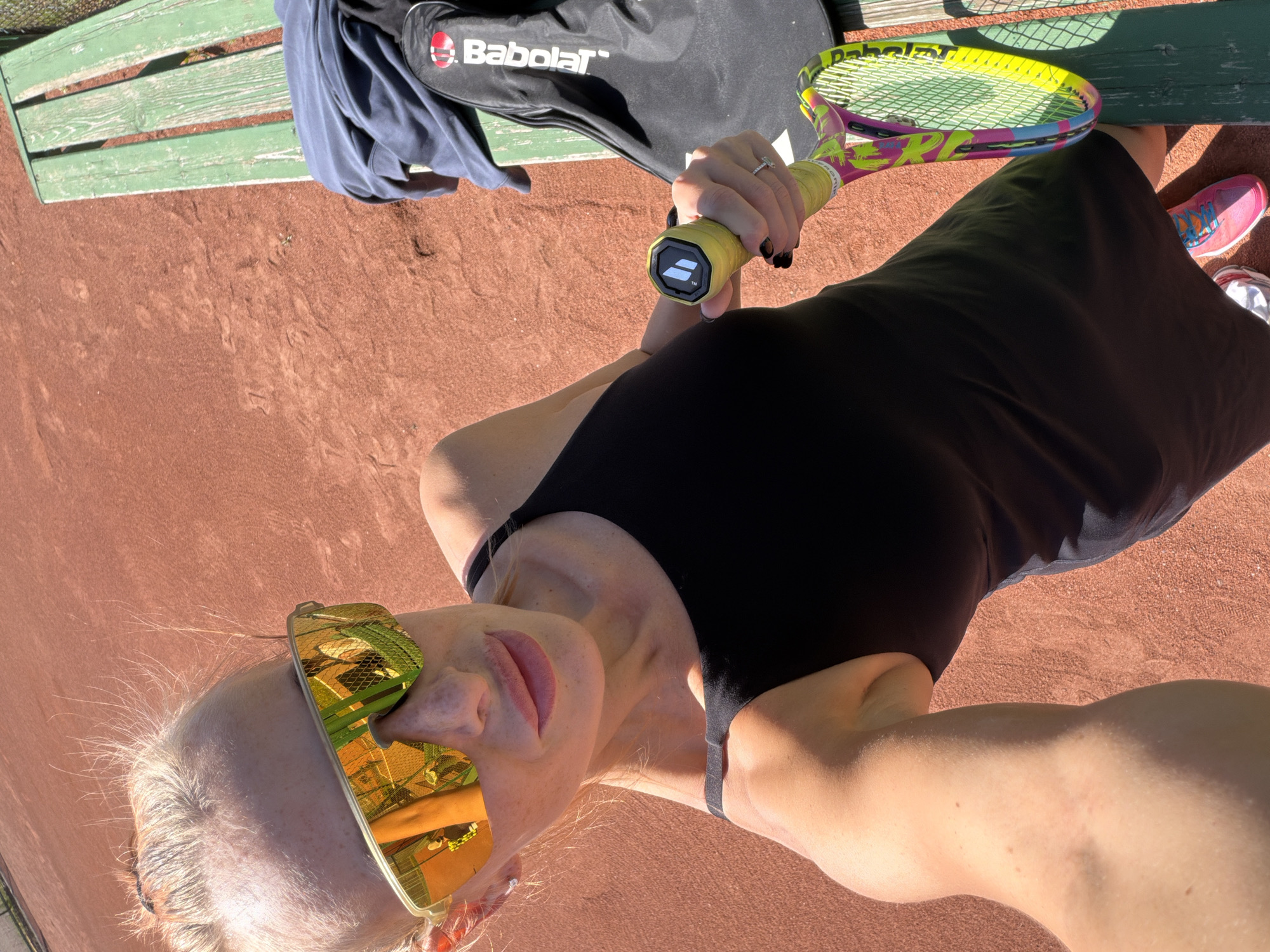 The image features a woman standing on a reddish-brown clay tennis court in Krasnoyarsk, Russia, under bright daylight. She is positioned as if looking slightly upward, with an overhead perspective. The woman has light-colored hair and wears a black sleeveless top or dress. She sports distinctive gold-mirrored sunglasses and a ring on her left hand.
She is holding a vibrant tennis racket, predominantly pink, yellow, and green, with the word "PURE" clearly visible on its frame. The butt cap of the racket handle also displays the Babolat logo. To her left, resting on a weathered green wooden bench, are a black tennis bag prominently featuring the "Babolat" brand name in white letters, and a navy blue piece of clothing. Parts of pink and white athletic shoes are visible near her feet.
The strong, direct sunlight indicates a clear, sunny day, casting distinct shadows on the court. The clay surface shows typical scuff marks and disturbances, suggesting recent play or an active tennis environment. The scene captures a moment of rest or a posed shot on a tennis court, combining sports equipment with a stylish, activewear aesthetic.