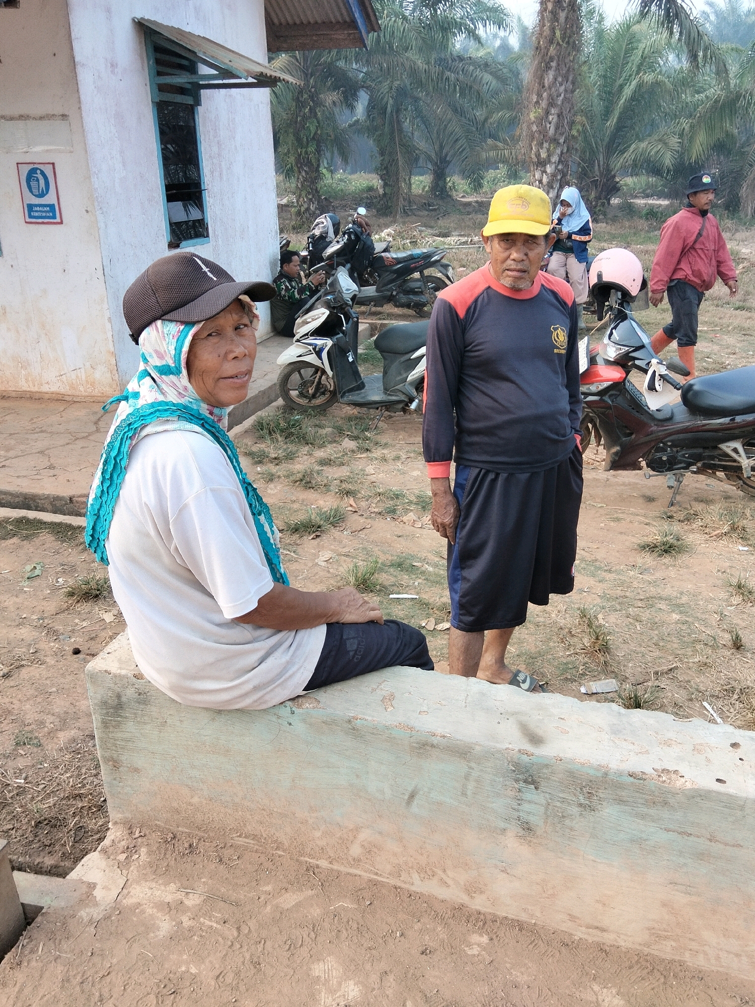 In Teluk Tenggirik, Indonesia, a woman wearing a white t-shirt, a hijab with blue and green accents, and a dark baseball cap sits on a light blue concrete bench. To her right stands a man in a long-sleeved dark blue shirt with orange accents at the collar and cuffs, dark blue shorts with a light blue stripe, and a yellow baseball cap. He is also wearing flip-flops. Behind them, several motorcycles are parked, and palm trees are visible in the background. A building with a light-colored wall and a corrugated metal roof is to the left of the frame, featuring a window with blue trim and a sign with a trash can icon and the text "JANGAN KEBERSIHAN" (Keep Clean). Another man in a red jacket and dark pants, wearing orange boots, walks away from the camera in the background to the right. A woman in a light blue hijab and light-colored pants is also visible in the background. The ground is covered with dirt and dry grass.