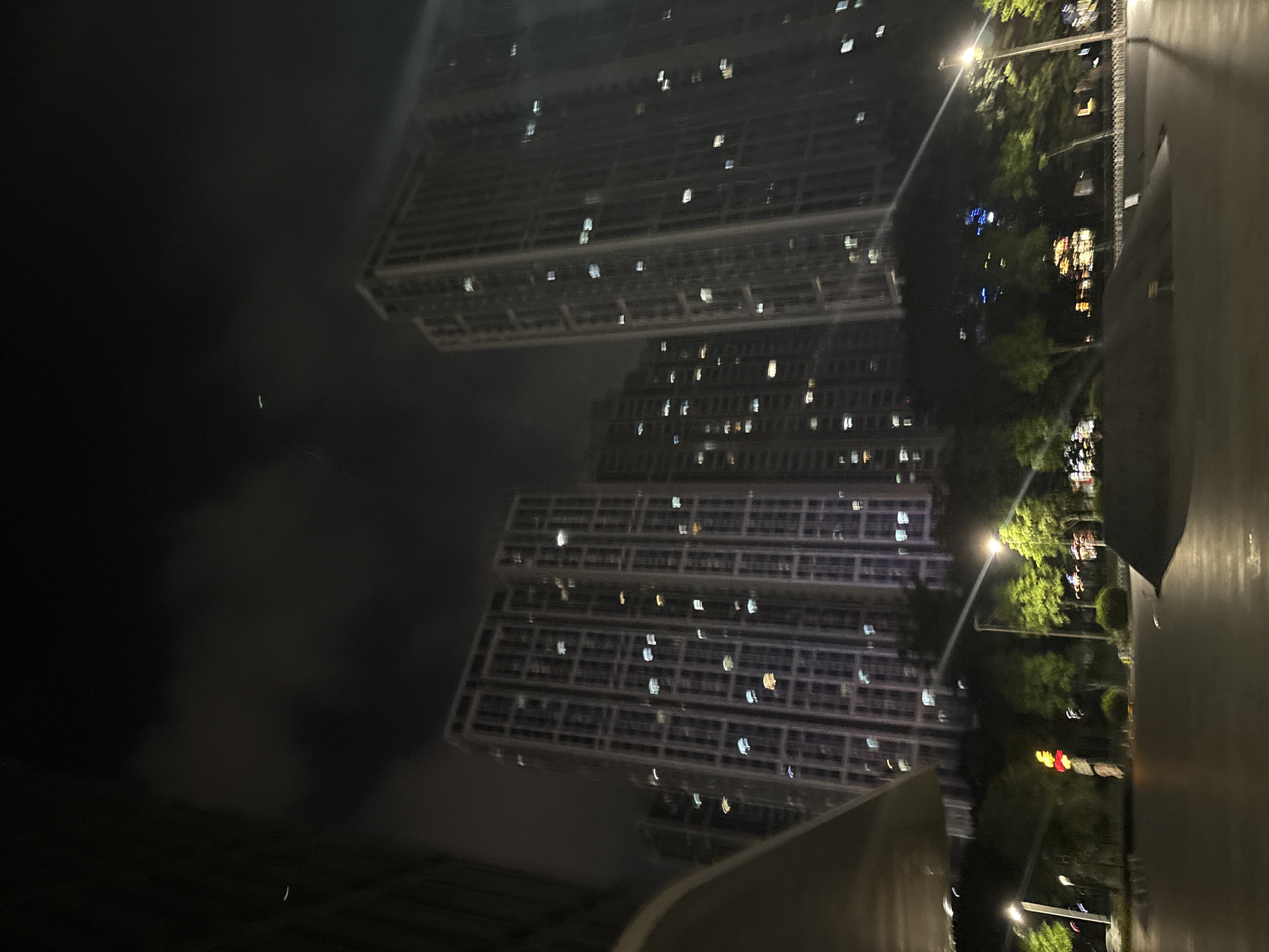This is a nighttime photograph taken in Haizhu District, China, featuring a modern urban scene. The primary subjects are two large, multi-story apartment buildings, their facades illuminated by countless glowing windows. The buildings are tall and rectangular, with a grid-like pattern of balconies or recessed windows. The lighting suggests it is nighttime, with the darkness of the sky contrasting with the artificial lights from the buildings and streetlights.
In the foreground and to the right, there is a wet, reflective surface, possibly a wet road or pavement, indicating recent rain or damp conditions. Trees with lush green foliage are visible, their leaves illuminated by streetlights, casting a soft glow. A streetlamp is prominently featured, its light beam cutting through the darkness. Further into the background, more lights from other buildings and possibly a traffic signal with red and yellow lights are discernible, suggesting an active urban environment.
The image is slightly blurred, giving a dreamy or impressionistic quality. There are no discernible people or specific activities captured. The overall mood is tranquil and slightly atmospheric, characteristic of a city at night after a rain. No visible text is present in the image.