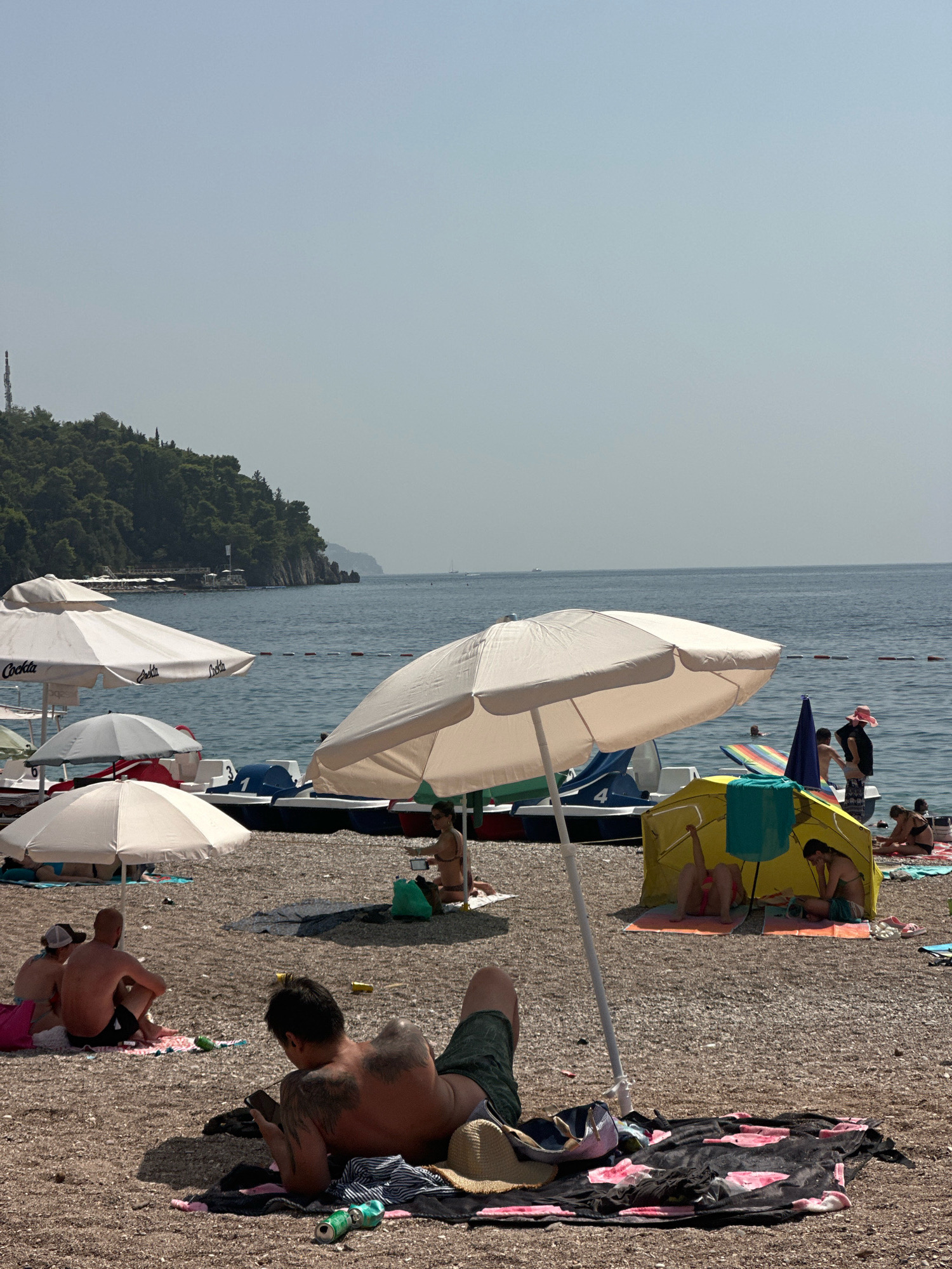 The image depicts a crowded pebble beach in Budva, Montenegro, on a clear, sunny day. The sea is calm and a light blue, with a rope barrier in the water marking a swimming area. In the foreground, a man with prominent tattoos is lying on his back, looking at his phone. He is on a beach towel with a straw hat and two cans of drink nearby. Several other people are scattered across the beach, some sitting, some lying down, and some under beach umbrellas.
In the middle ground, a yellow beach tent is set up with people inside. Various beach umbrellas, mostly white, are spread across the shore. Behind the umbrellas, several pedal boats are lined up, some with people in them. The backdrop of the beach features a lush green, hilly coastline with trees and buildings visible on the slopes. The overall atmosphere is one of leisure and relaxation during a summer day at the beach.