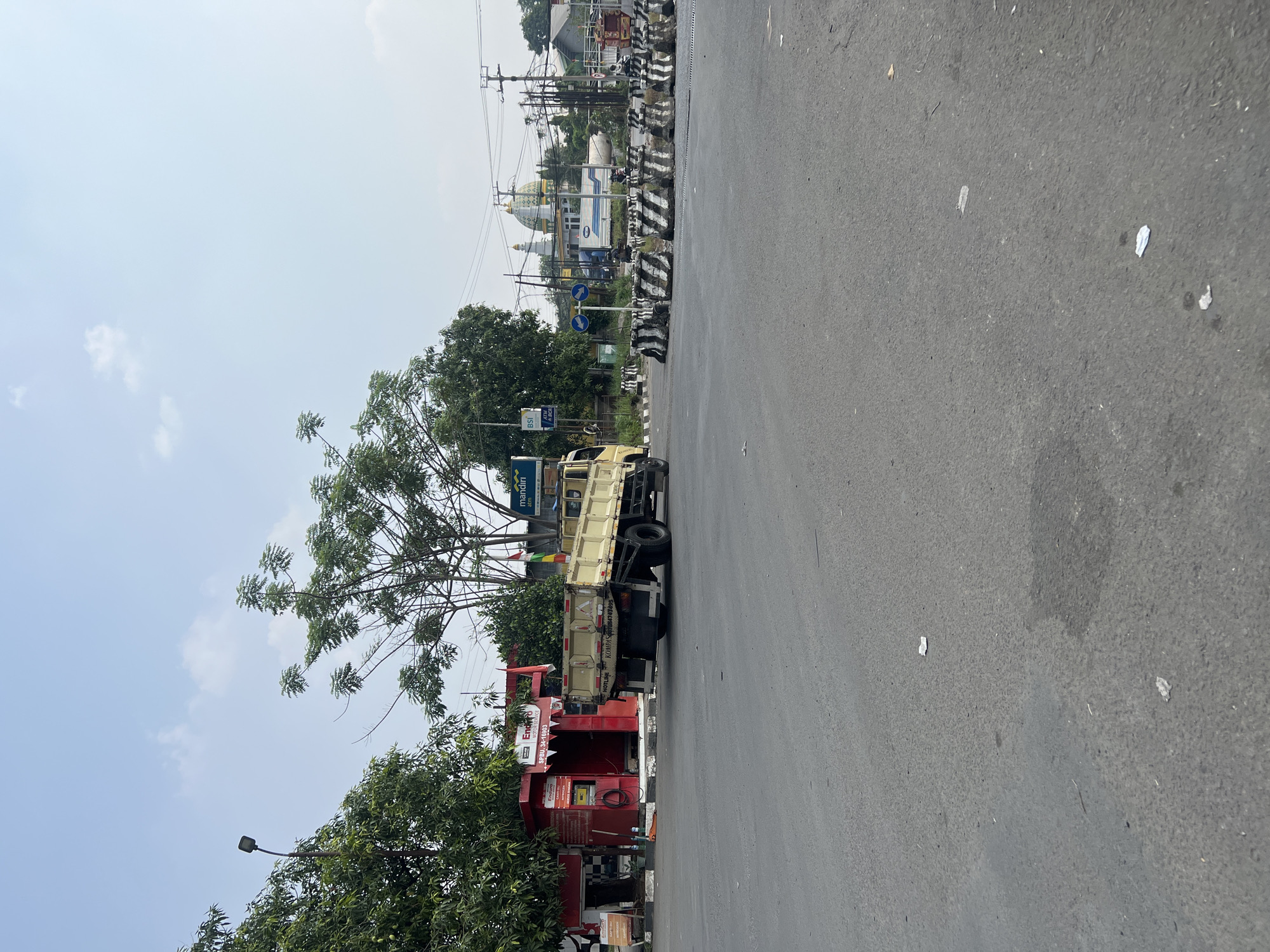 The image depicts a street scene in Gunung Putri, Indonesia. In the foreground, a two-lane asphalt road is visible, with scattered pieces of paper on the surface. A yellow flatbed truck with a beige cargo box is positioned on the right lane, facing away from the viewer and towards a road closure. The truck has black tires and a license plate that is difficult to read.
The road is lined with a series of concrete barriers, alternating black and white, indicating a partial road closure or a traffic diversion. Beyond the barriers, several trees with green foliage are present. To the left of the truck, a small red structure, possibly a kiosk or a small shop, with a white roof and "Enduro" branding is visible. A red sign with white text is attached to the front of this structure.
In the background, there are more trees and buildings. A prominent building with a green dome and white minarets, resembling a mosque, is situated in the distance. A white truck with "PERTAMINA" branding is parked behind the barriers. Power lines and poles are also visible throughout the scene. The sky is clear blue with some scattered clouds, suggesting a bright, sunny day. There are no people clearly visible in the image.