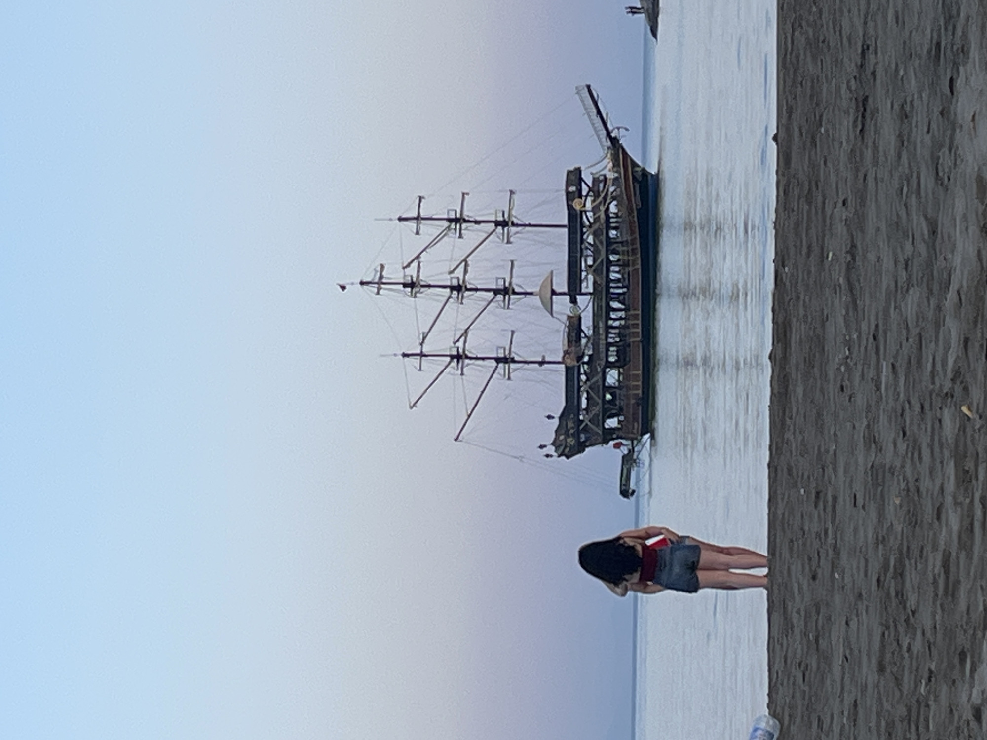 A wide shot captures a woman on a beach in Alanya, Turkey, with a docked pirate ship as the backdrop. The woman, dressed in a red top and a denim skirt, faces the ship on the water's edge. The ship has tall masts and dark wood. The right side of the shot shows the coarse sand of the beach. The sky transitions from light blue to a dusky purple. Another figure is visible in the far distance on the water.