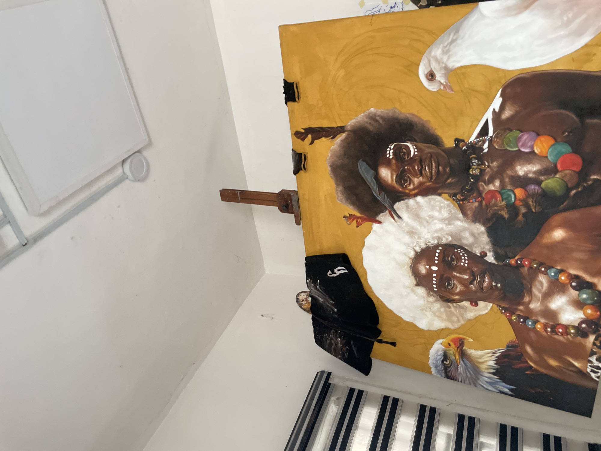 The image shows a painting displayed on an easel in a room with white walls. The painting depicts two dark-skinned individuals wearing traditional attire, including colorful beaded necklaces and face paint. One figure has an afro hairstyle, while the other has a large white hairstyle. A white dove is positioned above the first figure, and an eagle is below the second figure. The background of the painting is a yellow hue. A black cloth or garment hangs near the bottom of the painting. The room appears to be indoors. No specific location within Ado, Nigeria is discernable from the image.