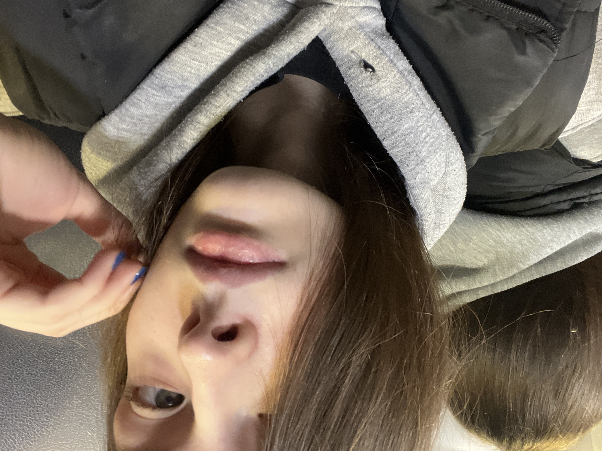 A selfie-style close-up image shows a person's face from an unusual, tilted angle, appearing almost upside down. The person has fair skin and brown hair falling around their face. Their lips are slightly parted. One eye is clearly visible, appearing brown. A hand with blue nail polish is near their ear. They are wearing a black jacket with a gray hooded sweatshirt underneath. The background is out of focus and indistinct. The location cannot be determined as Saint Petersburg, Russia based on the image alone.