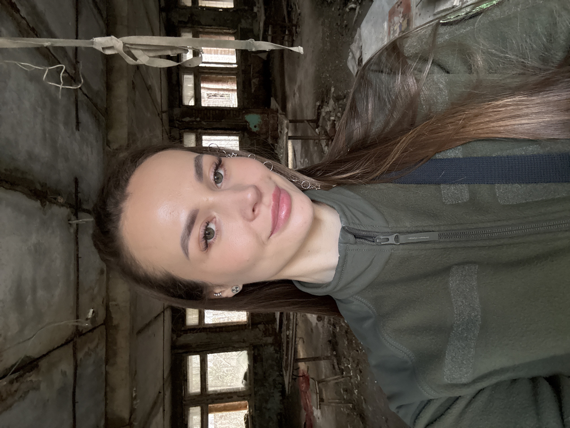 A selfie of a woman in a green fleece jacket, taken indoors in a deteriorated building. The background shows rows of window frames without glass. The concrete walls and ceiling are heavily damaged and worn, with exposed pipes and other building materials. The building appears to be located in a state of extreme disrepair and abandonment. Given the indication of Prypiat, Ukraine, the location could be related to the aftermath of the Chernobyl disaster.