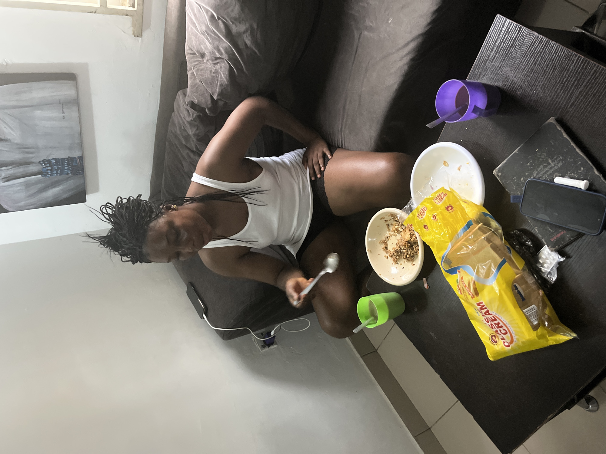 A Black woman with braided hair is sitting on a brown sofa in a dimly lit room, eating from a bowl. She is wearing a white tank top and black shorts. On a black coffee table in front of her are a bag of bread, two cups of drinks, a bowl of food, and a mobile phone. A painting hangs on the wall behind her. The scene appears to be set in an apartment in Abuja, Nigeria.