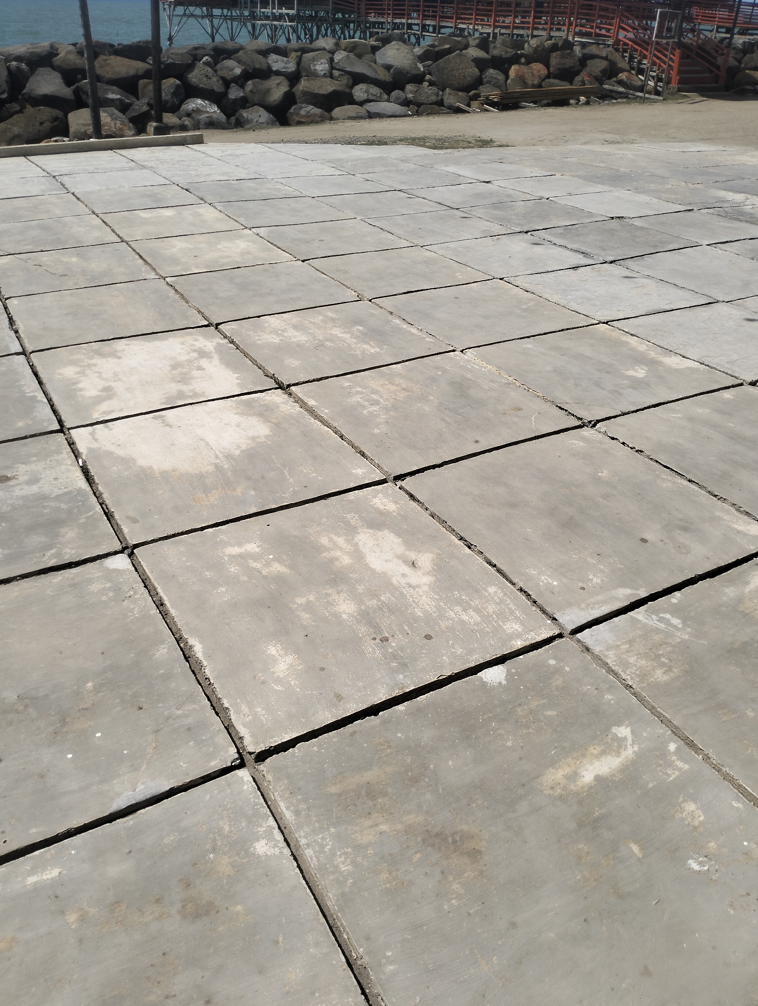 The image shows a paved area composed of large, square concrete tiles. The tiles are laid out in a grid pattern and exhibit signs of wear, with some discoloration and chips along the edges. The paving appears to be outdoors, possibly near a waterfront, as evidenced by the presence of a rocky embankment and a body of water in the background. Beyond the rocks, a pier or walkway structure with wooden elements and metal supports is visible. The scene suggests a coastal or harbor setting. The sky is bright, indicating daytime, and the lighting casts clear shadows, suggesting sunny weather. The overall impression is one of an open, public space with a utilitarian design. No people are visible in the image, and there is no discernable text. The location is identified as Lancok, Indonesia.