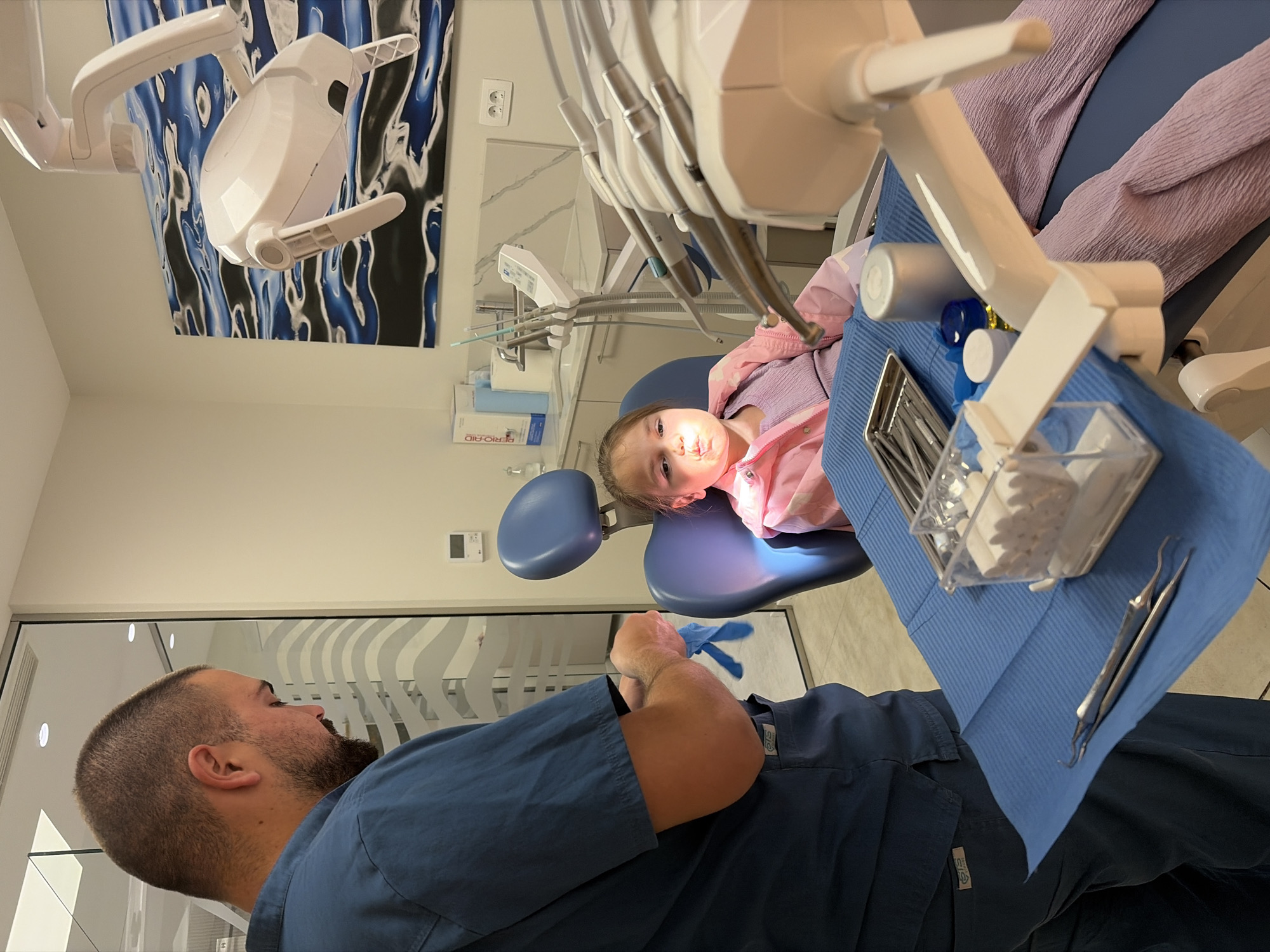 A dentist's office scene featuring a dentist and a young patient. The dentist, a man with a beard and short-cropped hair, is wearing blue scrubs. He is standing near the dental chair. A young girl in a pink jacket is seated in the dental chair, positioned under the dental light. The dental chair is covered with blue coverings and contains dental instruments. The room is well-lit, and there's a decorative painting on the wall. The location is the city of Budva, Montenegro.