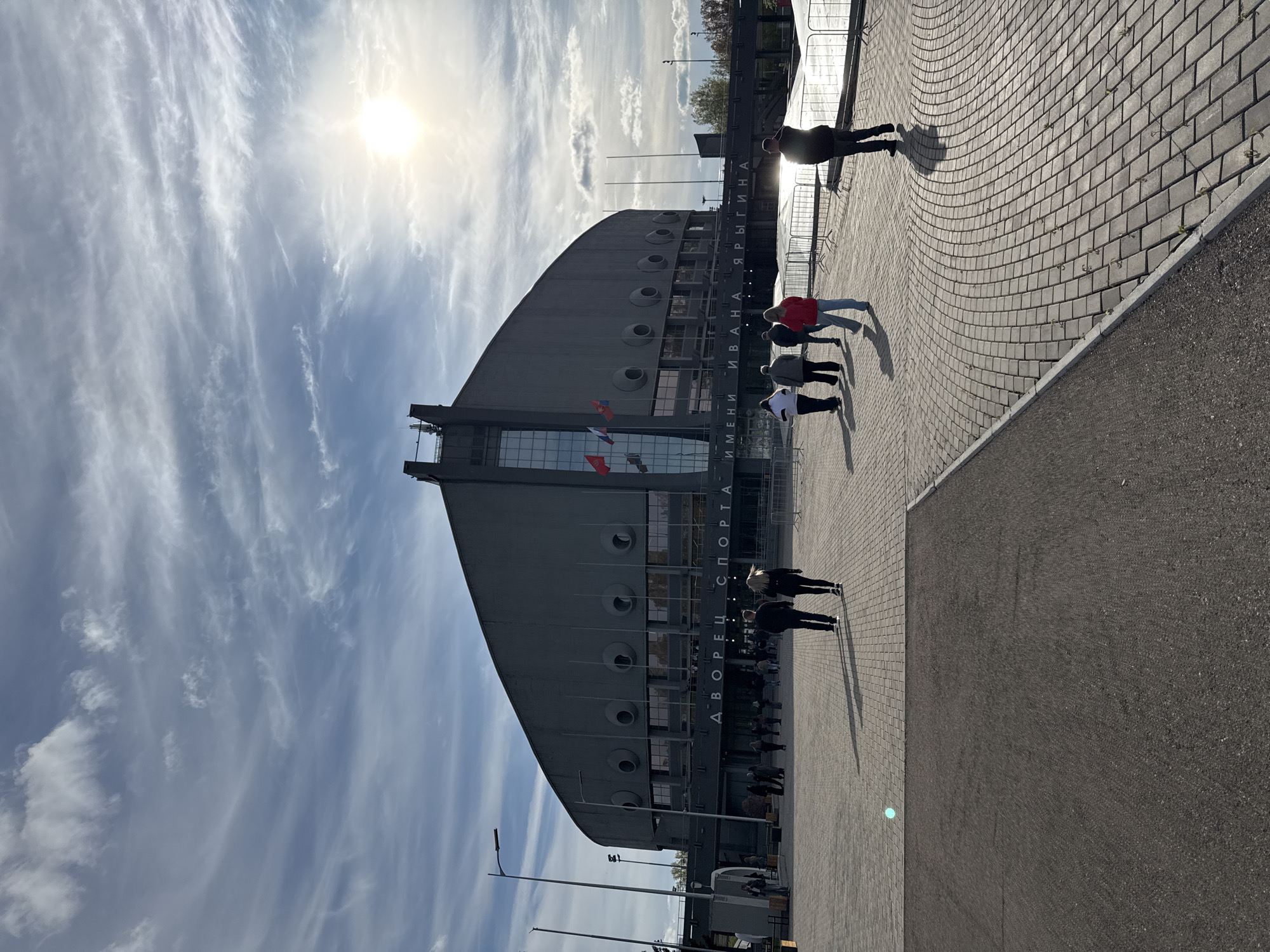 The image depicts the Ivan Yarygin Sports Palace in Krasnoyarsk, Russia, on a sunny day with a partly cloudy sky. The large, gray building, with its distinctive curved roof and circular window-like openings, dominates the scene. A group of people are walking on the paved walkway in front of the palace. One person is ahead of the group. The walkway is constructed of interlocking pavers and abuts a wider expanse of asphalt. The sun is bright and casts strong shadows. Flags are visible on the facade of the sports palace. Text in Russian is displayed on the front of the building.