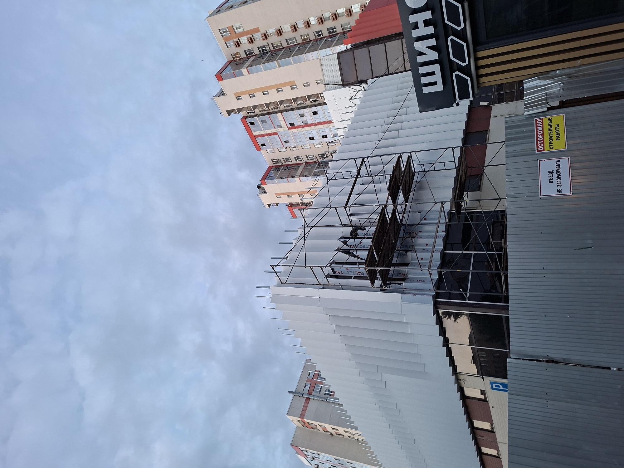 The image shows a construction site in Kazan, Russia, featuring a partially completed building with a white, corrugated facade and scaffolding. The building's design includes repeating vertical panels, giving it a modern aesthetic. In the background, taller residential buildings with red and beige accents are visible. A sign on the scaffolding reads "ALTEC" and another partially visible sign in the foreground displays the Russian word "ШИНО" which likely refers to a tire shop. Two warning signs are posted on a metal fence: one reads "ВЪЕЗД НЕ ЗАГОРАЖИВАТЬ" (Do not block entry), and the other says "ОСТОРОЖНО! СТРОИТЕЛЬНЫЕ РАБОТЫ" (Caution! Construction work). The sky is overcast, suggesting a cloudy day. There are no visible people in the image.