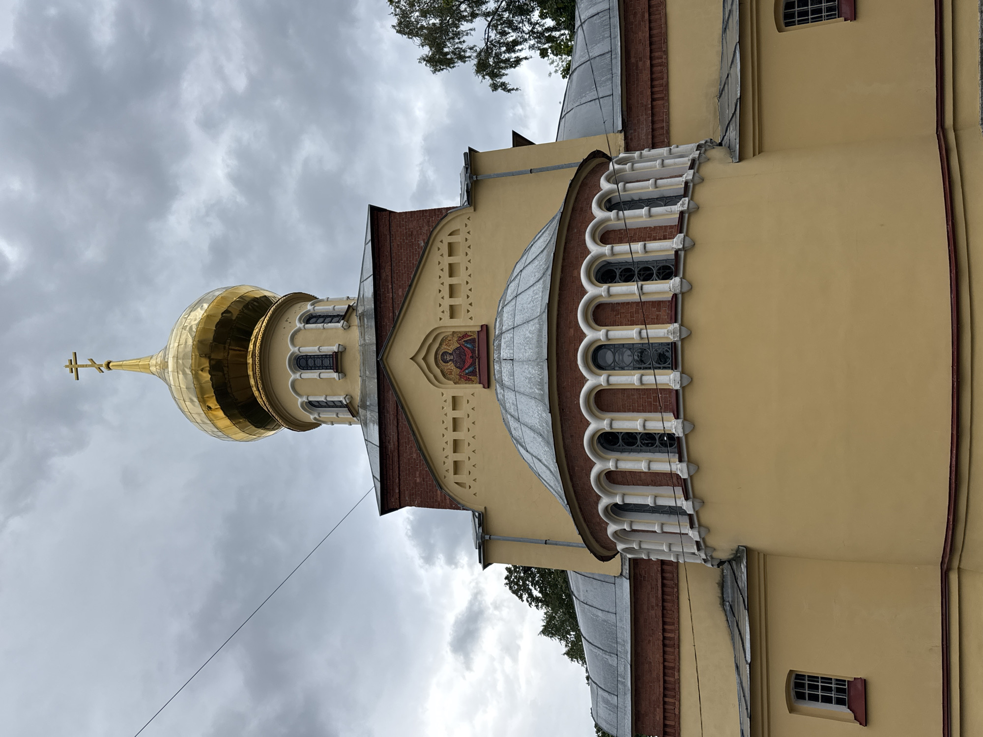 A low-angle, eye-level medium shot captures the facade of a yellow church with a golden dome under a cloudy sky in an unknown city and location. The building features red brick accents, arched windows, and a mosaic icon above a rounded bay with white columns. Trees are visible to the right of the church. A thin wire stretches across the sky in the upper left corner of the frame.