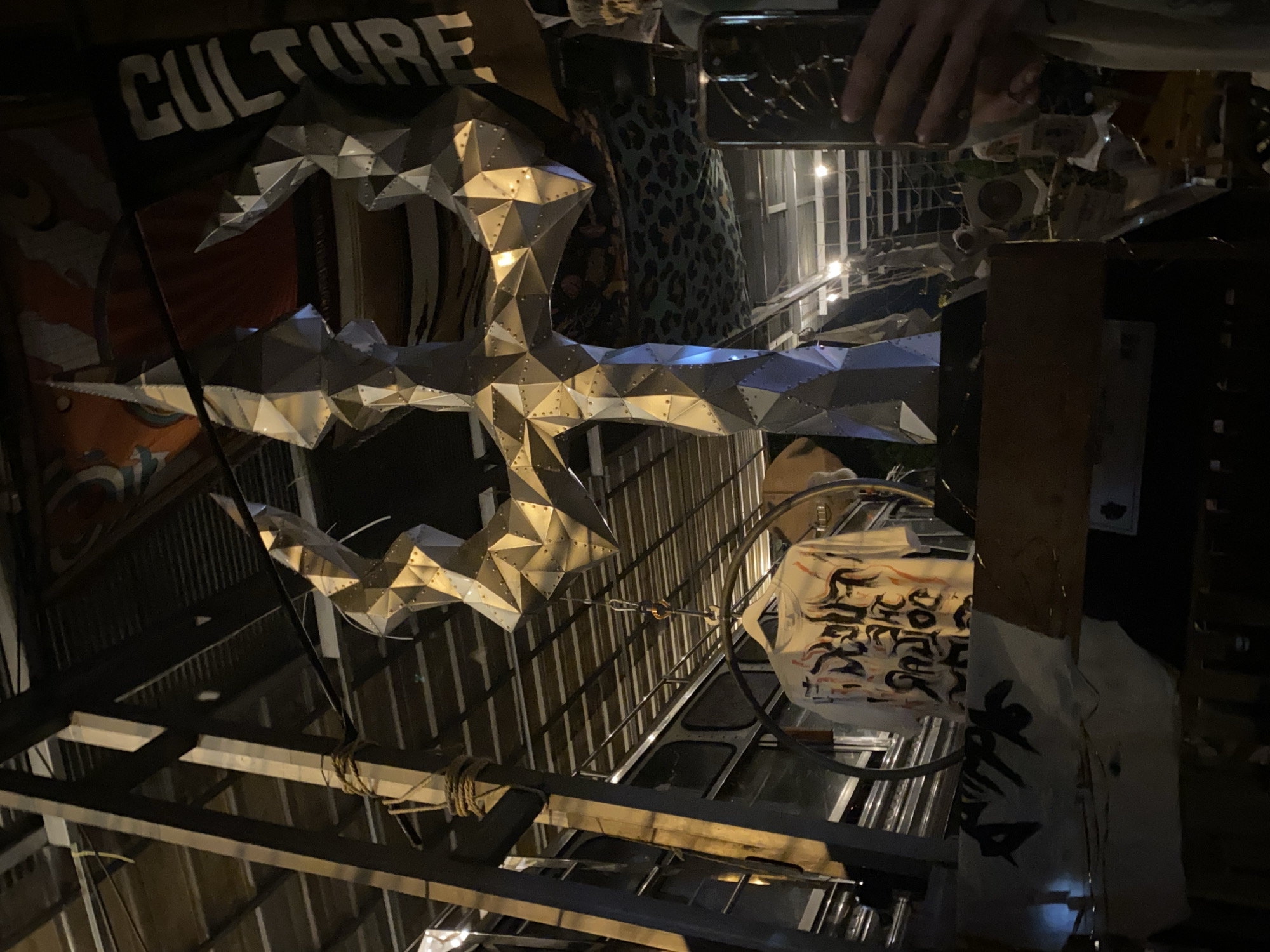 The image depicts an interior space with various objects and architectural elements. A large, silver, geometrically shaped object hangs prominently in the foreground. Behind it, a sign with the word "CULTURE" is partially visible. Other visible items include a rack, a printed bag, a metal cage, and various building materials and structures. A hand holding a mobile phone is visible in the top right corner, suggesting someone is taking a photo or video. Based on the prompt, this scene is located in Baan Nai Suan, Thailand.
