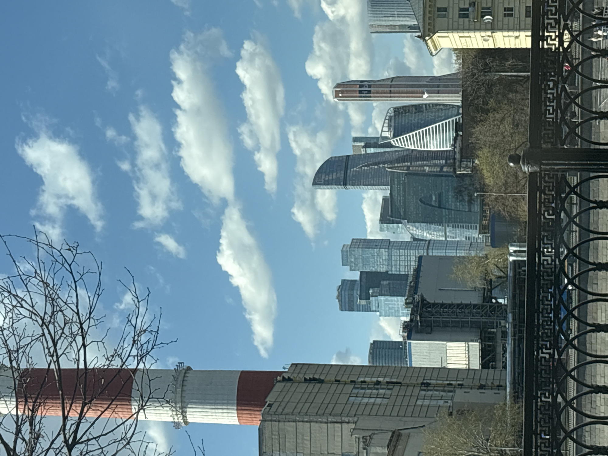 The image captures a view of the Moscow International Business Center (Moscow City) skyline in Moscow, Russia, under a partly cloudy sky. A tall, red and white striped industrial chimney is visible in the foreground, partially obscured by bare tree branches. Adjacent to the chimney is a building with a corrugated metal roof. A black, ornate metal railing runs along the right edge of the image, possibly part of a bridge or embankment. In the background, a cluster of modern skyscrapers, including the Evolution Tower and the Mercury City Tower, rise above the city. A building with a yellow facade and multiple windows can be seen behind the skyscrapers, possibly an older, Soviet-era structure. The sky is blue with elongated white clouds.