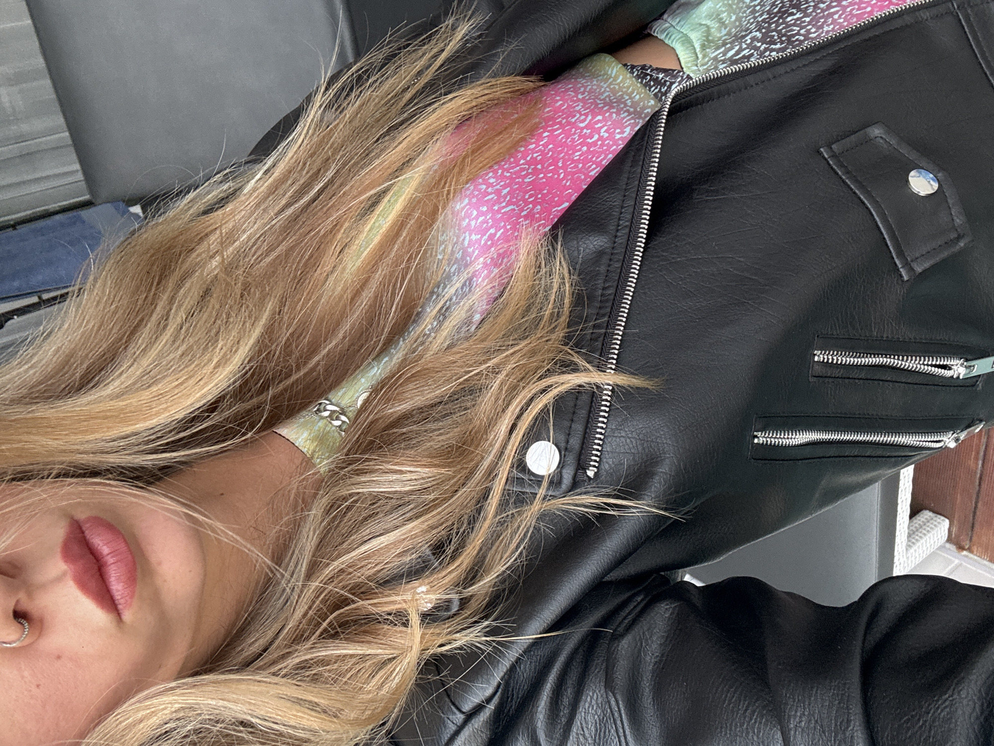 A person, partially visible from the chest up, is wearing a black faux leather moto jacket over a colorful, patterned top. The person has blonde hair and a nose ring. The background appears to be outdoors with a blurred dark gray surface and a hint of a white woven surface in the lower left corner. The image is a close-up shot, focusing on the person's attire and facial features.