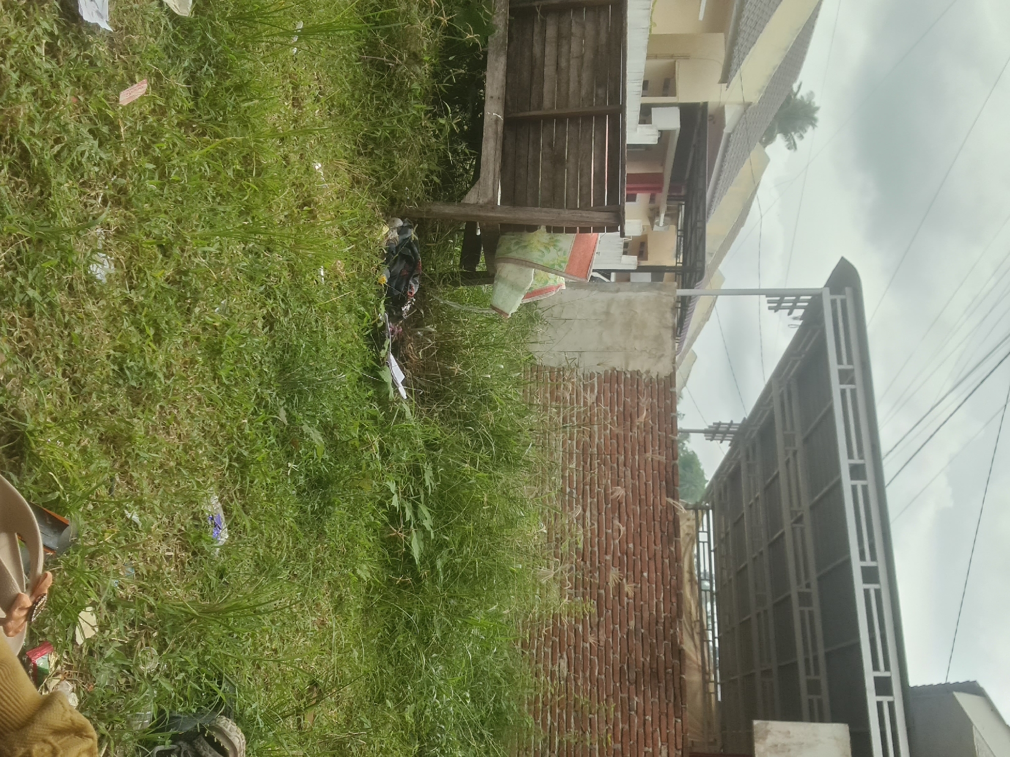 The image depicts an outdoor scene in Lhokseumawe, Indonesia, characterized by overgrown grass and debris. In the foreground, a wooden bench is positioned amidst the tall grass. Behind the bench, a pile of dark-colored clothing and other miscellaneous items can be seen. A light-colored bag with a floral pattern rests on the bench. Further back, a brick wall forms a backdrop, and beyond that, a modern building with a metal roof structure is visible. The sky is overcast, suggesting cloudy weather. The presence of the bench, discarded items, and untamed vegetation indicates a neglected or underutilized outdoor space, possibly a yard or a vacant lot.