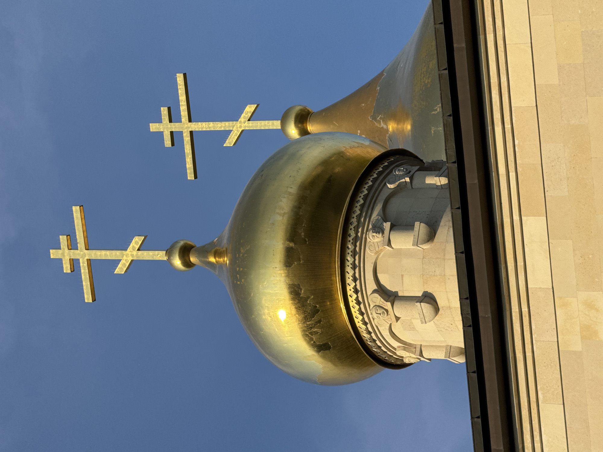 The image shows the golden domes of the Resurrection Cathedral in Kirishi, Russia, against a clear blue sky. The foreground features a close-up of two of these domes, topped with Orthodox crosses. The larger, left dome is highly reflective, catching the sunlight and showing some signs of wear. The dome to its right is partially obscured and also bears the golden cross. Beneath the domes, decorative stone elements with sculpted faces are visible. The lower portion of the image reveals the light-colored stone facade of the building, with a dark metal trim. The scene is static, with no people or animals present, focusing solely on the architecture of the cathedral.