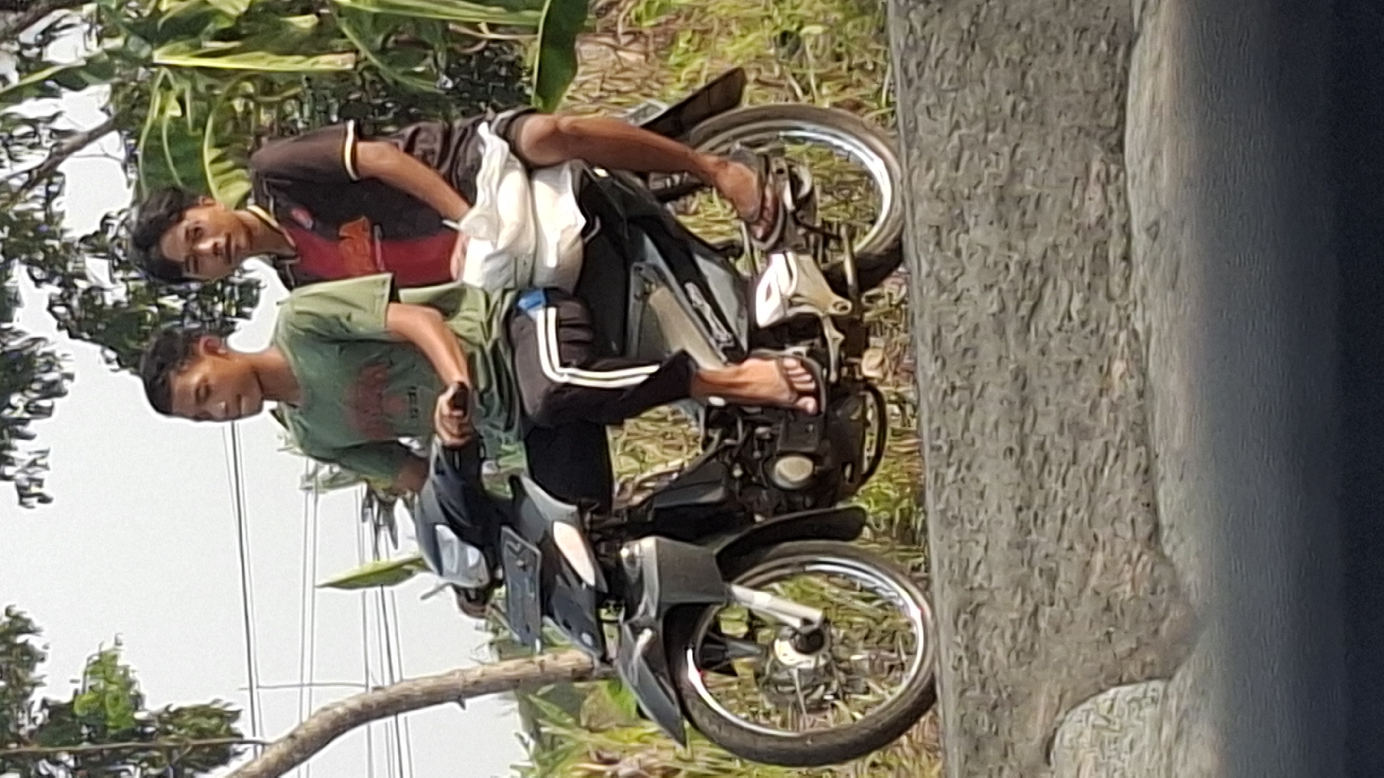 Two young males are riding a black motorcycle on a road. The rider in front, wearing a green t-shirt and dark pants with white stripes, has his hands on the handlebars. The passenger behind him, wearing a black and red t-shirt, is holding a white cloth. Both individuals are wearing flip-flops. The motorcycle is a scooter-style with a black front fairing and chrome accents on the wheels. They are positioned on a slightly elevated, textured grey surface, possibly a curb or a low wall. The background shows foliage, including green leaves and trees, with some power lines visible in the distance. The sky is overcast. The scene appears to be in a semi-rural or suburban area of Cibinong Hilir, Indonesia.