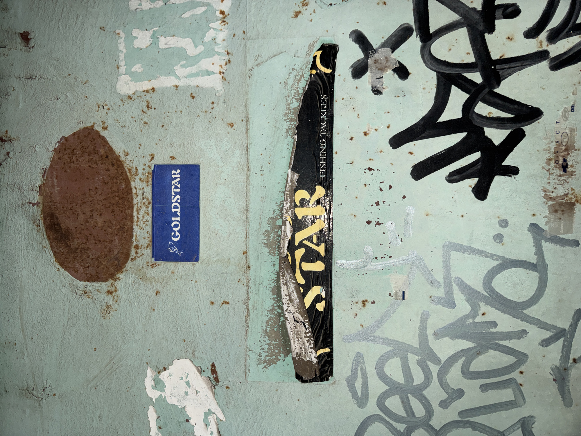 A close-up shot shows a section of a weathered wall in Sofia, Bulgaria. The wall's surface is a faded blue-green, with areas of peeling paint and rust. Affixed to the wall are several stickers and patches of paper. A blue sticker labeled "GOLDSTAR" is clearly visible. Adjacent to it is a rusted, oval-shaped stain. A black sticker with the word "STAR" in large gold letters and the phrase "FISHING TACKLES" is placed vertically. Black graffiti markings are also visible on the right side of the wall, adding to the urban decay aesthetic.