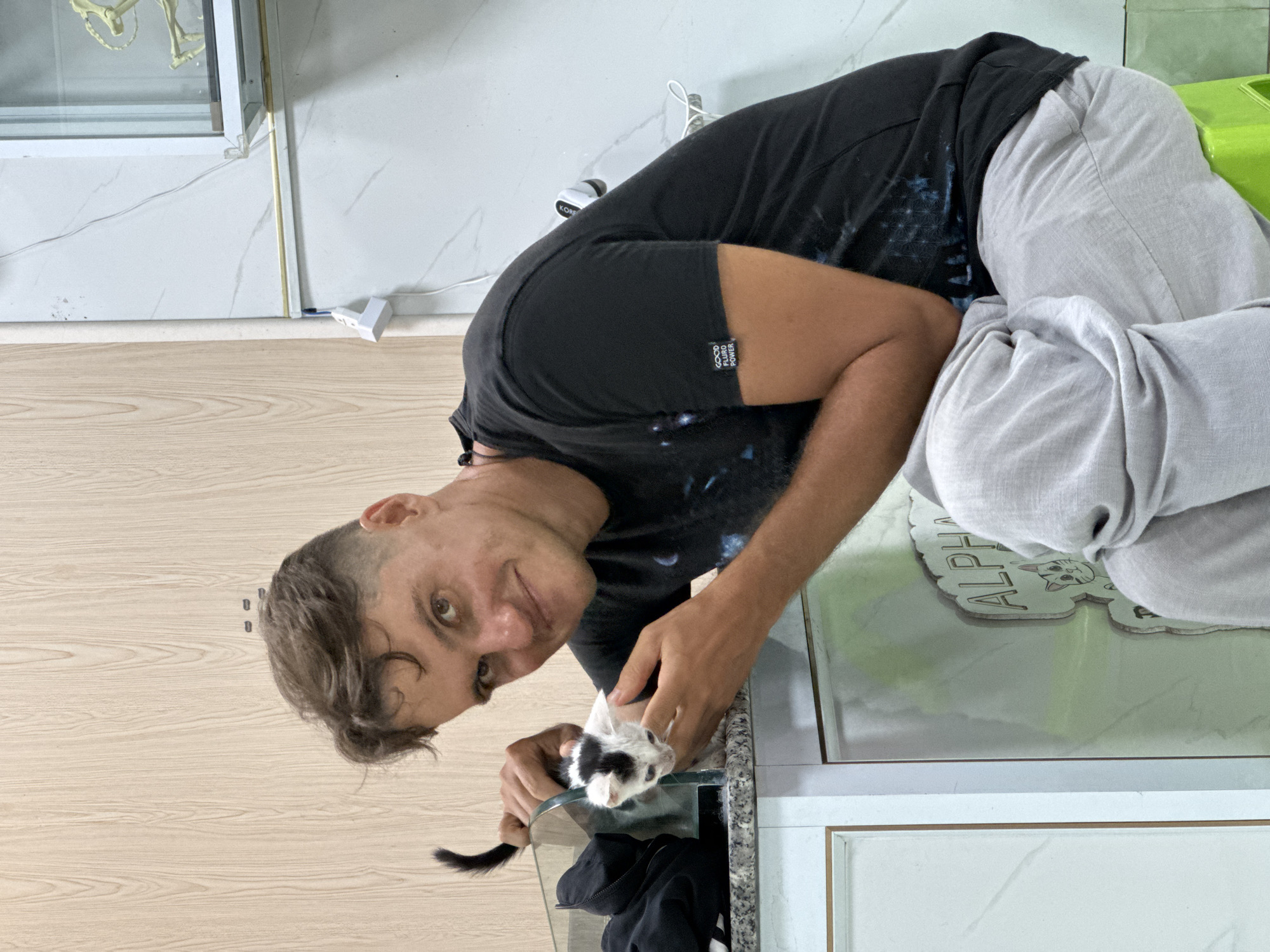 A man is sitting on a green stool in what appears to be a residential or commercial space. He is wearing a black t-shirt with a graphic print and light gray pants. His left arm is around a black and white kitten, which is perched on a glass surface. The kitten is looking down and to its left. Behind the man, there is a light-colored wooden wall and a tiled wall with some electrical outlets and wires. To the right, a glass-enclosed structure is visible. A sign with the word "ALPHA" and a cat illustration is also visible. The environment appears to be indoors.