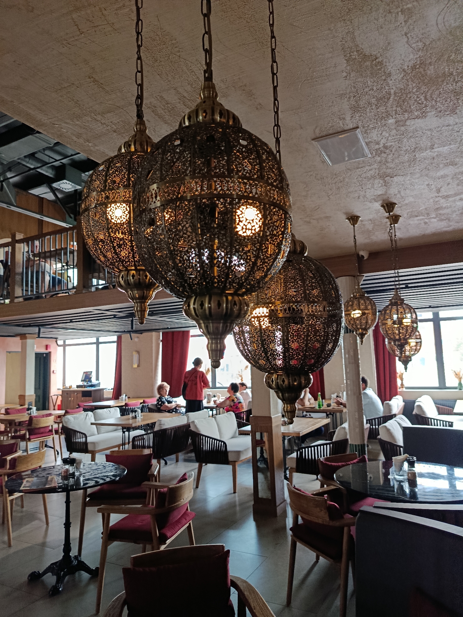 The image depicts the interior of a restaurant or cafe, likely located in Saratov, Russia, characterized by its ornate decor and relaxed ambiance. Dominating the foreground are three large, intricately designed metal pendant lamps with a Moroccan or Middle Eastern influence, hanging from the ceiling by chains. These lamps cast a warm, ambient light through their patterned metalwork.
The restaurant features a multi-level design. The main dining area, visible in the lower portion of the image, is furnished with tables and chairs. Several tables are round with dark, reflective tops and intricate black metal bases, accompanied by chairs upholstered in deep red fabric. Other seating arrangements include white upholstered sofas and benches, suggesting a comfortable and modern seating concept.
In the mid-ground, several patrons are seated at tables, engaged in dining or conversation. A man in a red shirt appears to be interacting with patrons near a window. Further back, there is a raised mezzanine level with a railing, offering a different vantage point of the restaurant. Large windows line the back wall, allowing natural light to enter and providing a glimpse of the exterior, though the specific view is unclear. Red curtains are visible, adding a touch of color and elegance to the space.
The overall environment suggests a well-lit, possibly upscale establishment with a focus on atmospheric lighting and comfortable seating. The combination of decorative elements, lighting, and seating arrangement creates an inviting and stylish dining space.