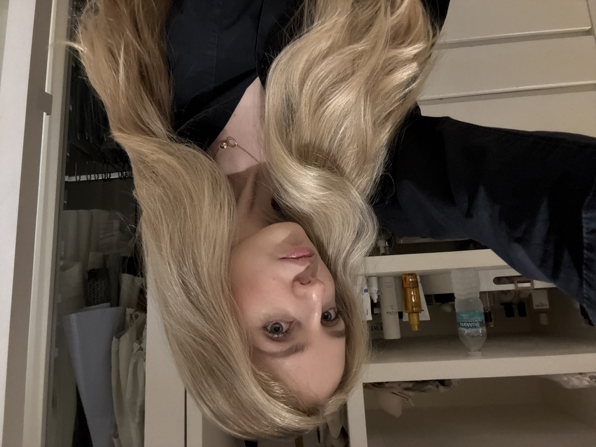 A woman with long, blonde, wavy hair is facing the camera. She is wearing a black shirt and a delicate necklace. Her gaze is direct and her expression is neutral.
Behind her is a white closet. The closet's contents, partially visible, include clothing, a shelf with bottles, and a water bottle labeled "Santa Maria."
The location of the scene is inside, likely in Mexico City, Mexico, based on the brand of the water bottle.