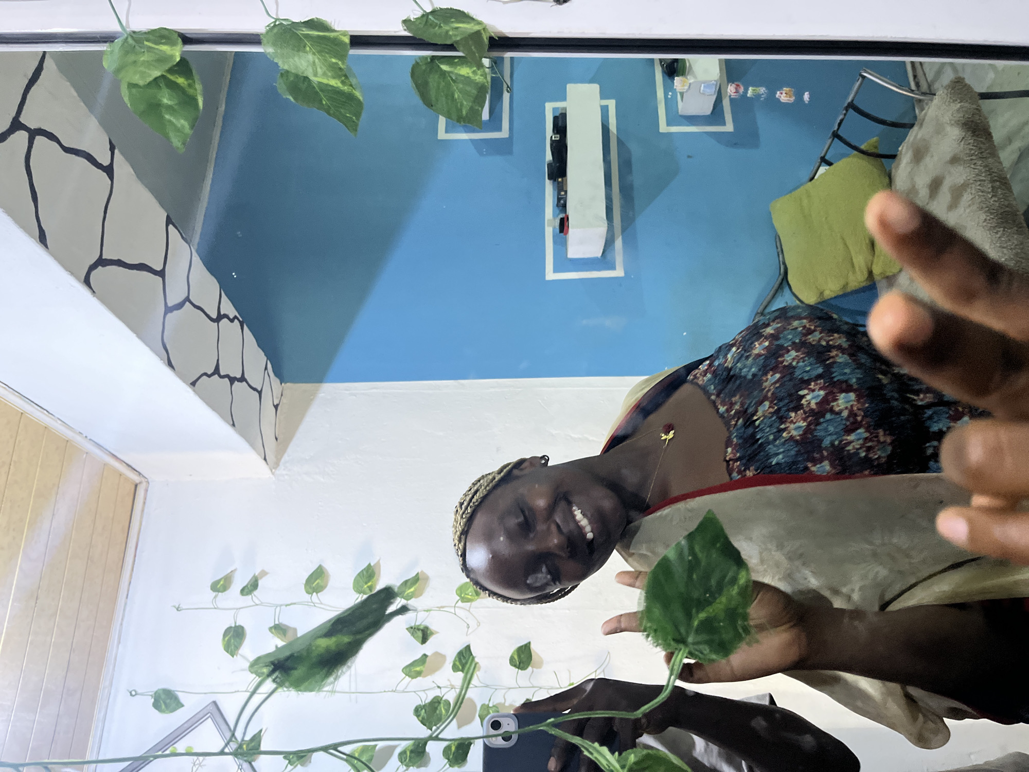 A person is indoors. The person is making a peace sign. There are green plants. The walls are white with black accents and solid blue.