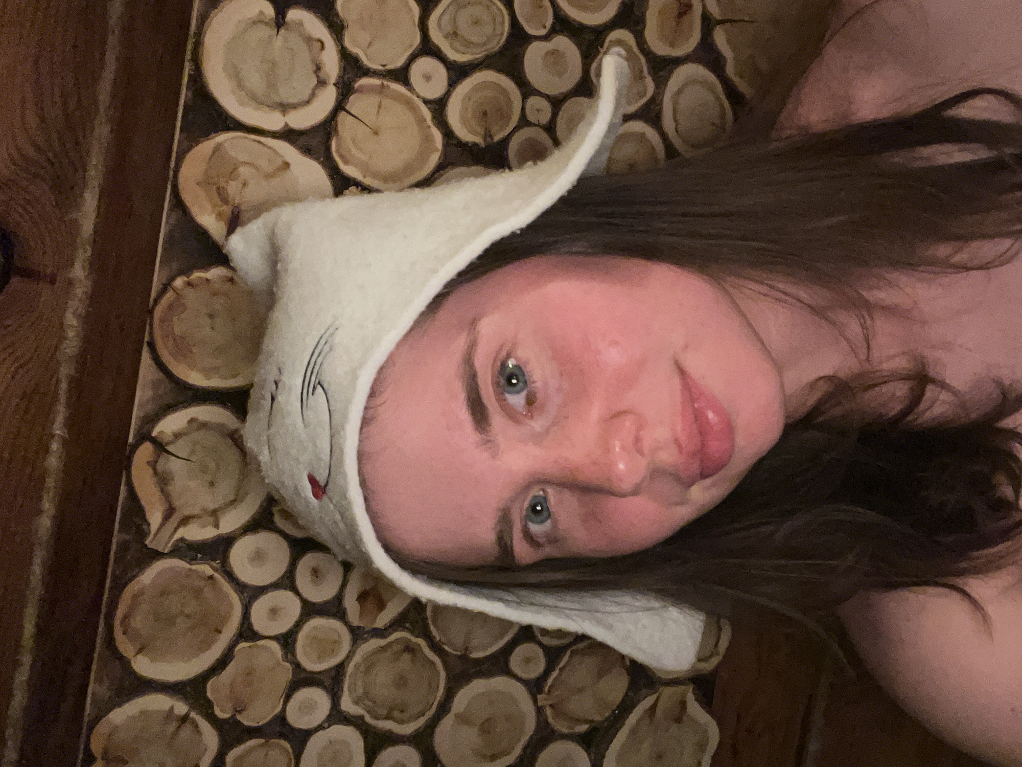 A fair-skinned woman with long brown hair and blue eyes is lying on a wood surface. She is wearing a white felt sauna hat shaped like a cat. The wood surface appears to be composed of small, round wood slices. The image was taken indoors. It is unknown if the location is Chaikovsky, Russia.
