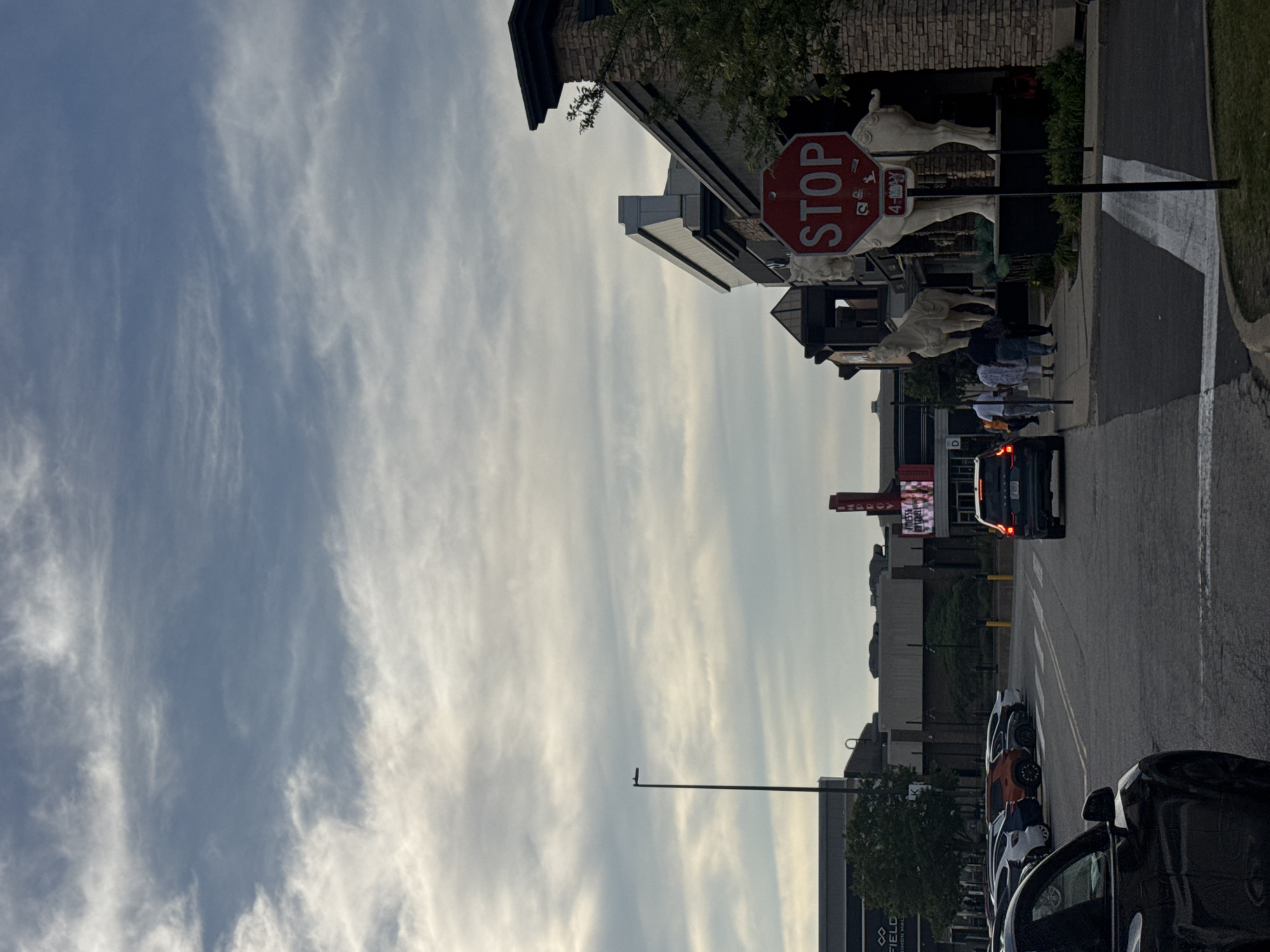 The image depicts a street scene in Schaumburg, Illinois, United States. The sky is predominantly visible, featuring wispy clouds against a light blue backdrop. On the right side of the frame, a street runs vertically with parked cars lining the edge. A stop sign is mounted near a building facade adorned with statues of horses. Pedestrians can be observed walking on the sidewalk. In the background, a car with its brake lights illuminated is visible, along with other commercial buildings and signage.