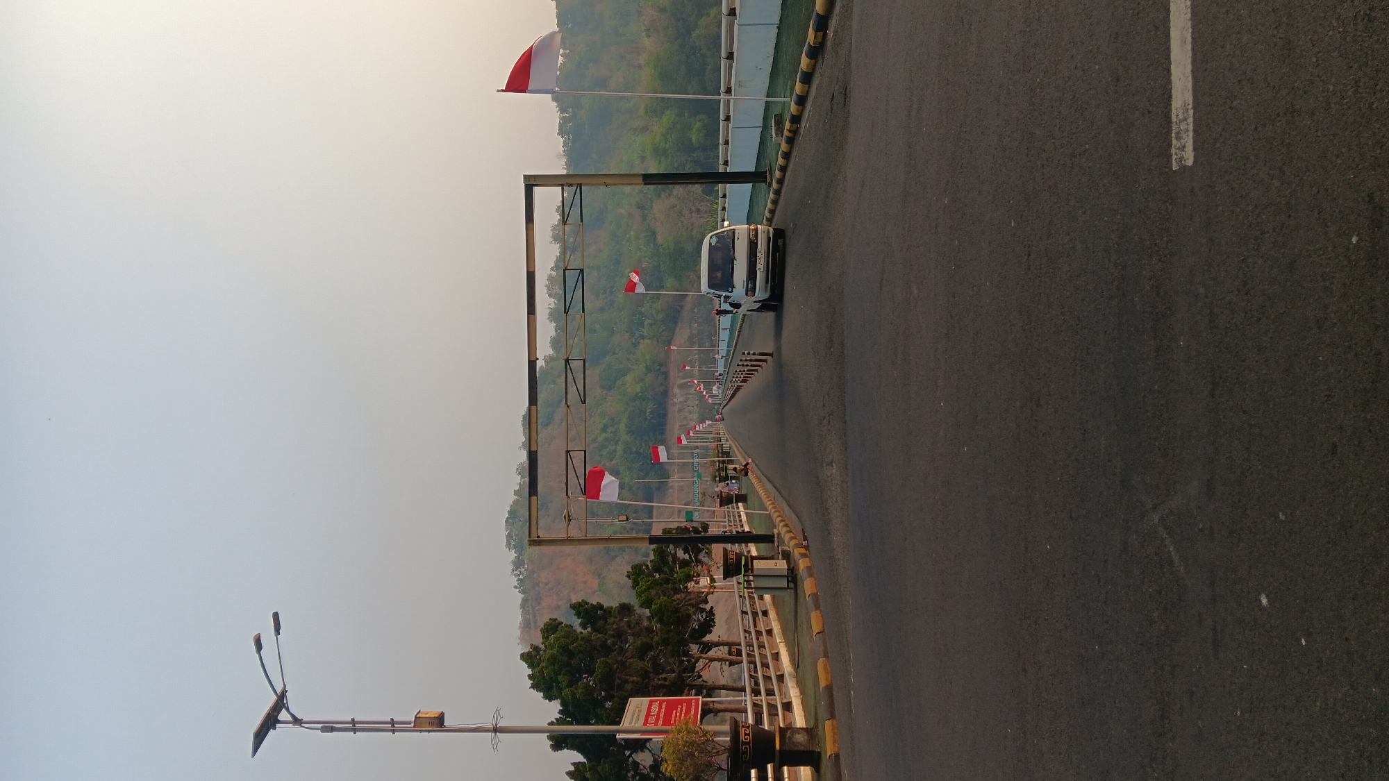 The image shows a road leading across a dam, likely the Jatiluhur Dam in Purwakarta, Indonesia, given the presence of Indonesian flags and the hilly terrain. The road is paved and appears to be open to vehicular traffic. On the left side of the road, there is a pedestrian walkway with railings, trees, planters, and a lamppost with a solar panel. A sign with red text is also visible on the left. Several Indonesian flags (red and white) are planted along the sides of the road, some on poles and others attached to an overhead structure. A white van is driving away from the camera on the right side of the road. In the background, there are green, tree-covered hills, and the sky is a hazy, light blue, suggesting a warm day or atmospheric conditions. A few people are visible sitting on the pedestrian walkway in the middle distance. The scene appears to be a public area, possibly a tourist or recreational spot, and the flags indicate a nationalistic or celebratory context.