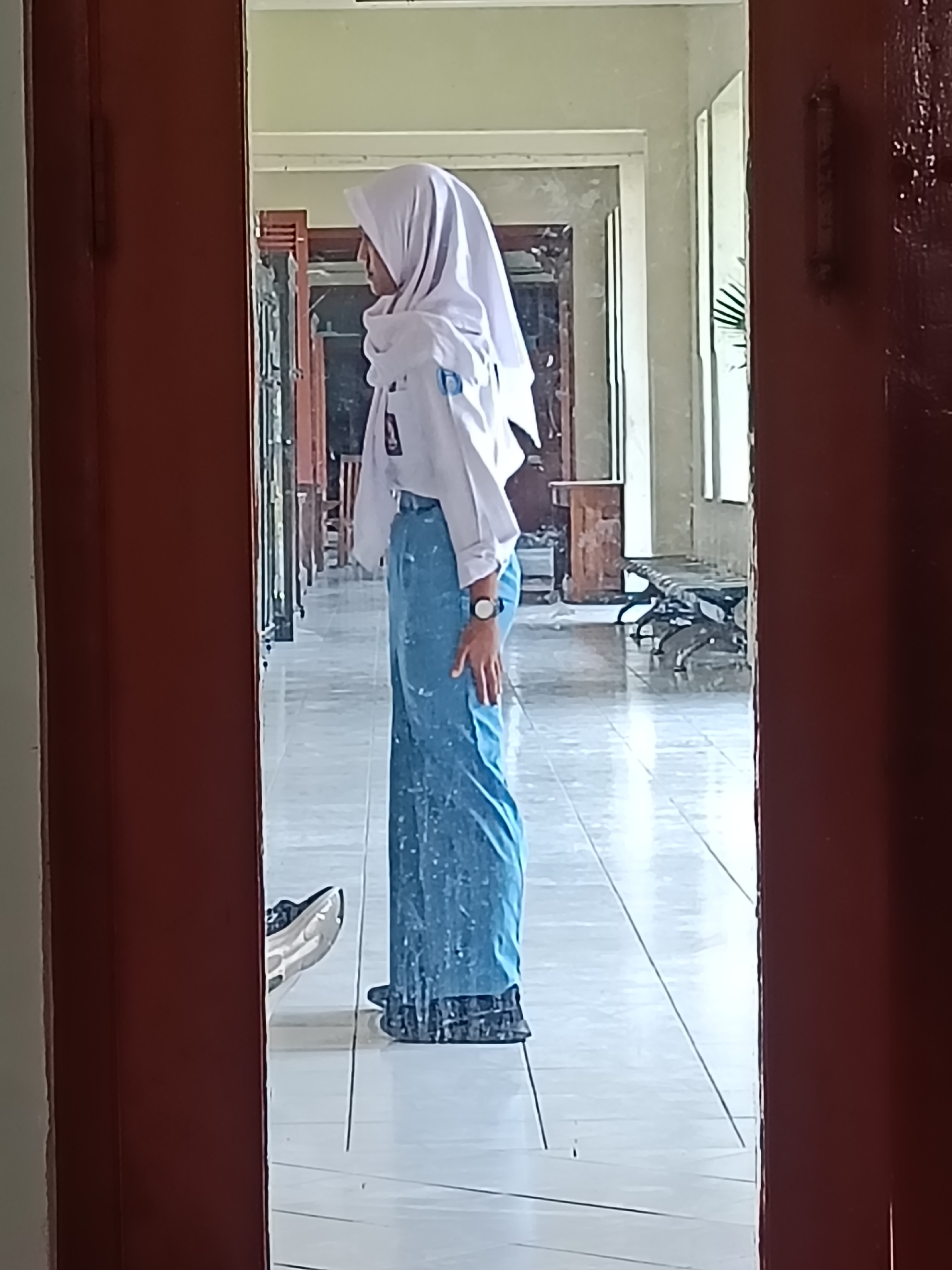 A young person wearing a white hijab and a white school uniform shirt with blue pants stands facing a mirror. The person's left side is visible, with their head turned slightly to the left. They appear to be in a hallway with a tiled floor and windows or doors on the right side. The reflection in the mirror shows the hallway extending into the background with benches and some lockers or cabinets. The scene is likely within a school building in Kediri City, Indonesia, given the context of the school uniform.