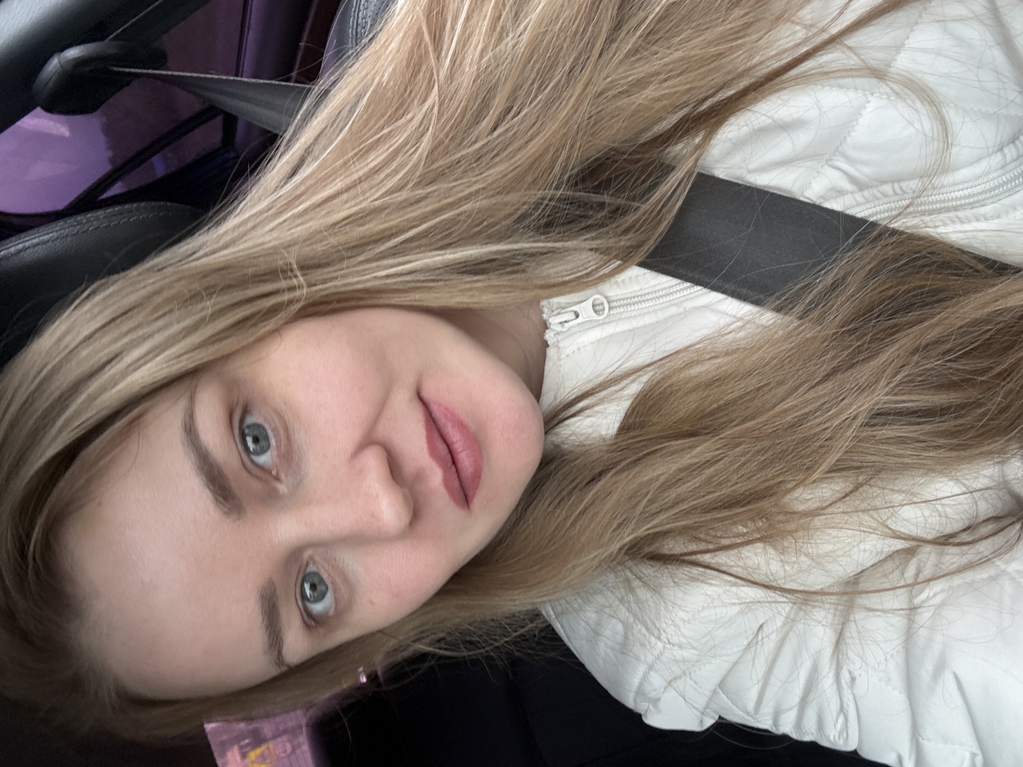 A young woman with long blonde hair and blue eyes is wearing a white quilted jacket and a black seatbelt. She appears to be in a car. It is not possible to verify the location.