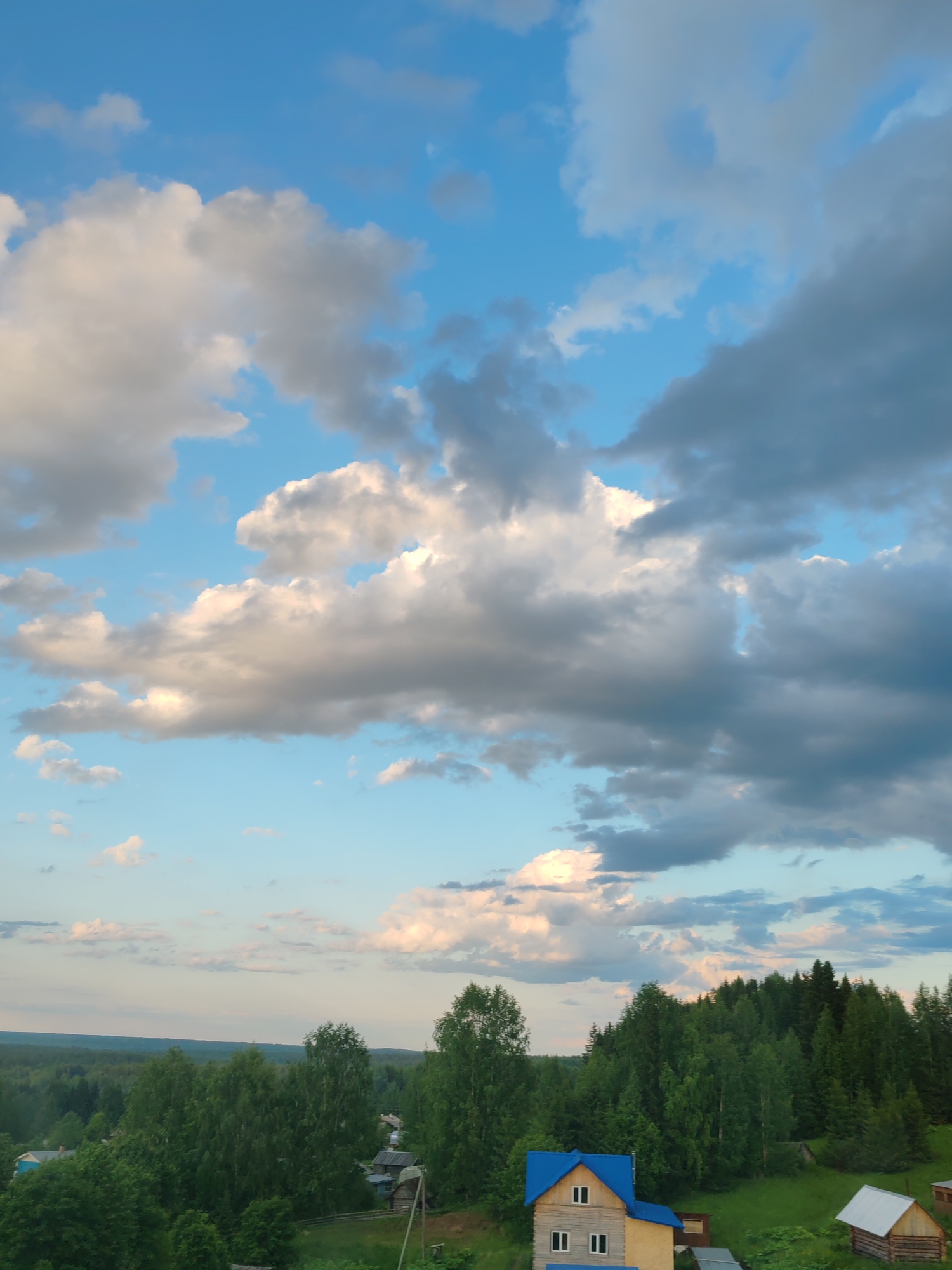 The image captures a wide view of a rural landscape under a dramatic sky filled with clouds. In the foreground, a cluster of trees frames the scene, with a bright blue-roofed house with yellow walls and white-trimmed windows prominently displayed on the right. To the right of this house, a smaller wooden structure with a white roof is visible. The middle ground features a dense forest of green trees that extends across the horizon, with glimpses of a few scattered houses and structures among the trees. The background is dominated by a vast expanse of sky, characterized by a mix of bright blue and large, textured clouds, some illuminated by sunlight. The overall impression is one of a peaceful, bucolic setting in the countryside. Based on the architecture and the surrounding vegetation, this scene could be located in a rural area such as Чухлэм, Russia, which is known for its picturesque natural beauty and traditional wooden houses. There are no people visible in the image.